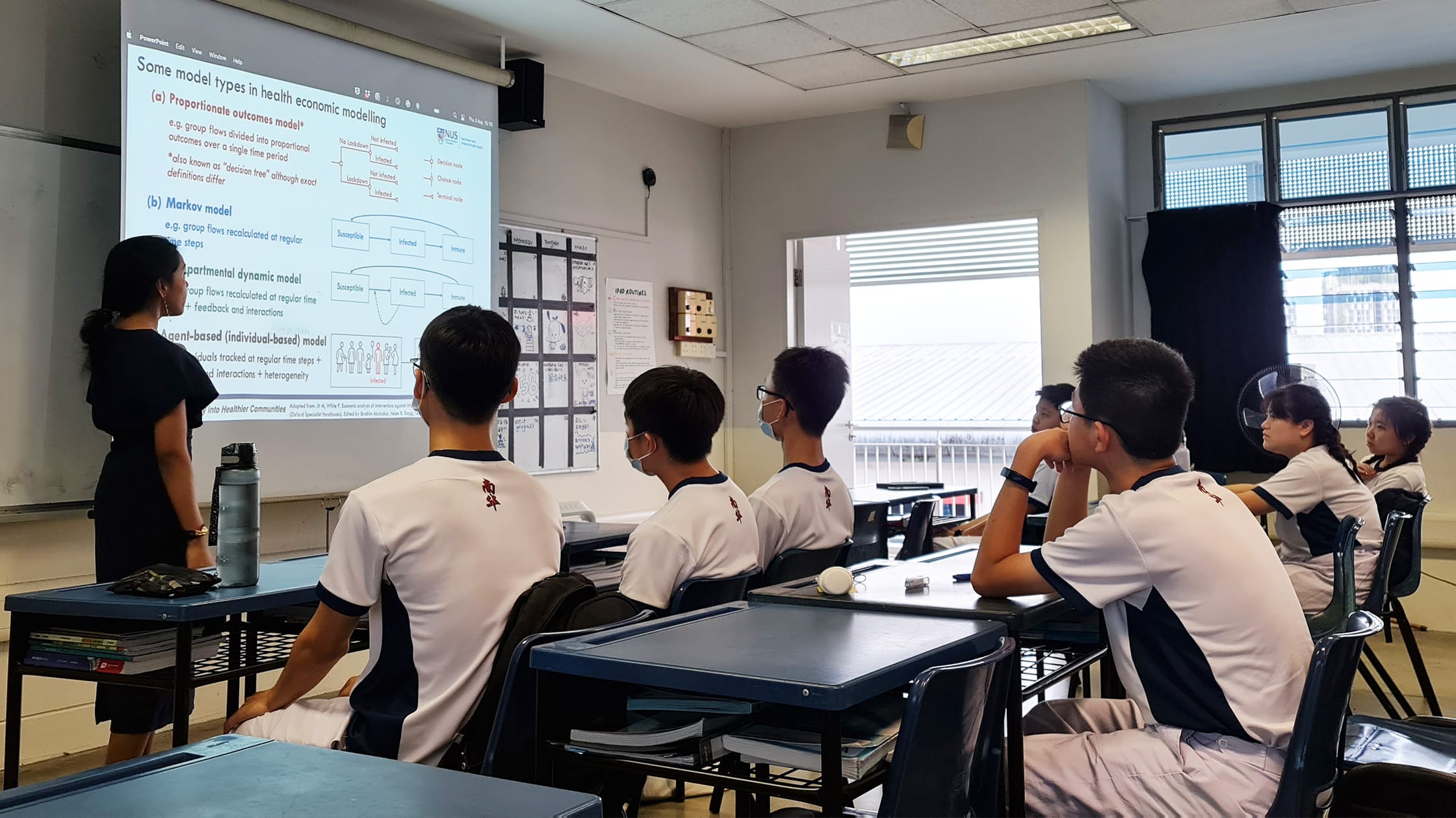 Students in white uniforms sit at desks facing a projected slide on health economic modelling presented by a teacher.