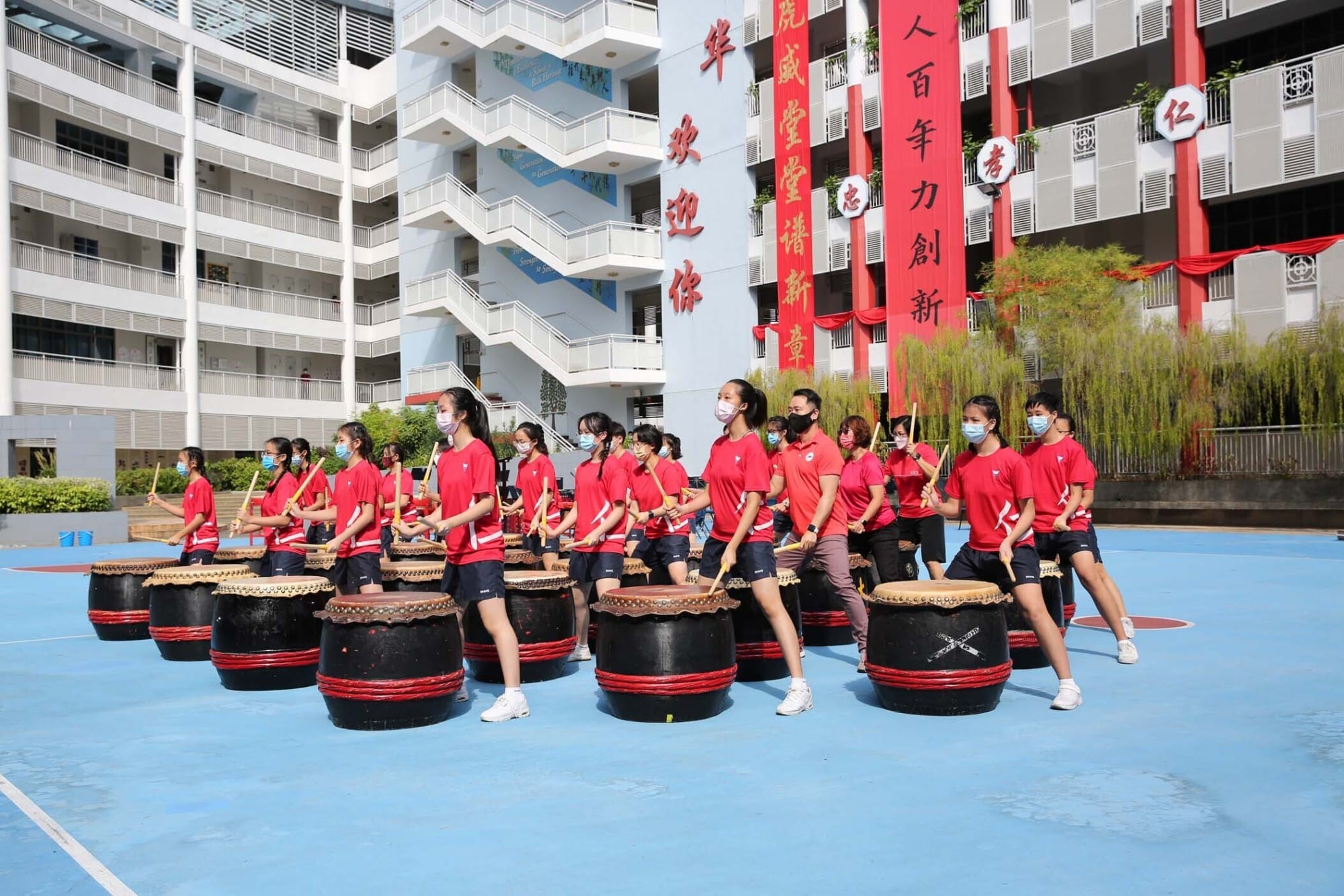 Students in red shirts and masks play large traditional Chinese drums outdoors in a school courtyard with Chinese banners.