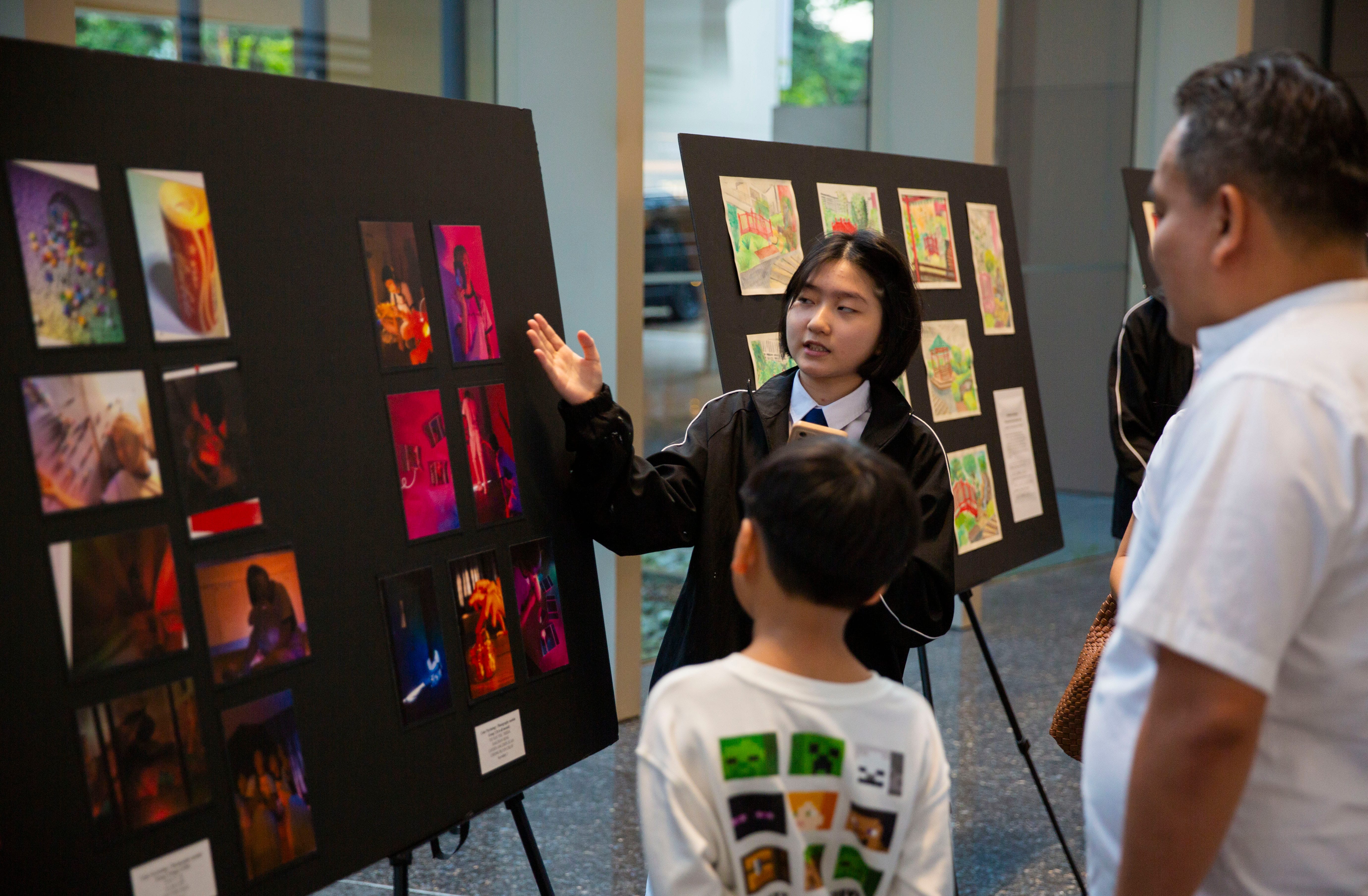 A young woman gestures toward display boards of colorful artwork while speaking to a child and adult at an art show.