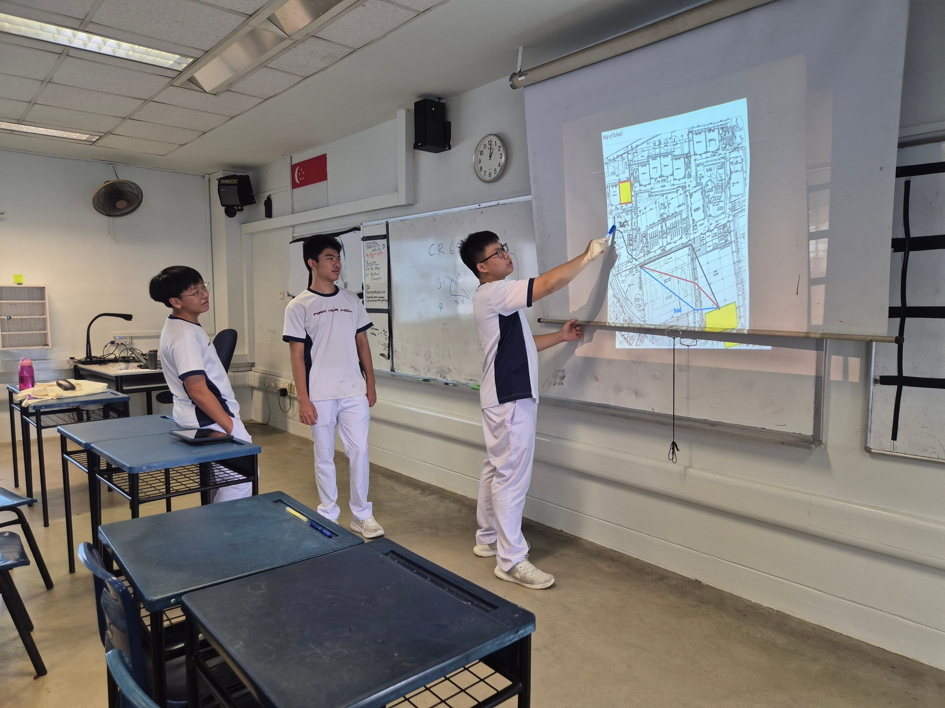 Three students in white uniforms stand in a classroom, one pointing at a projected map on a screen.