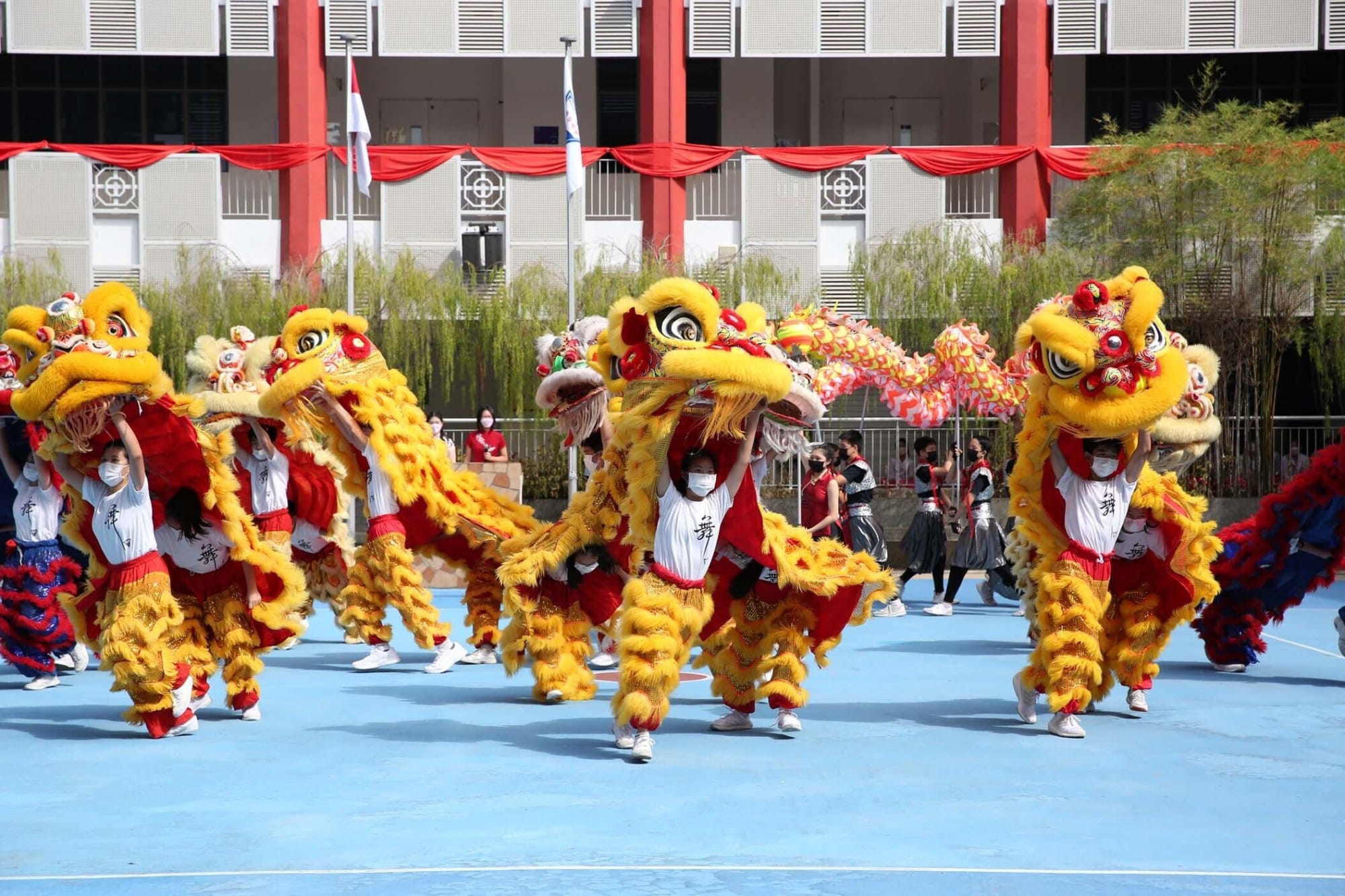 Performers in white and red costumes carry bright yellow and red lion dance puppets on a blue outdoor court.