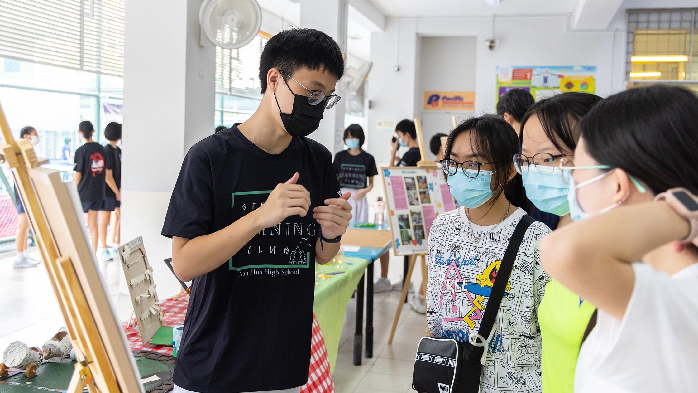 Students wearing face masks gather around display boards and easels at a school exhibition or fair.