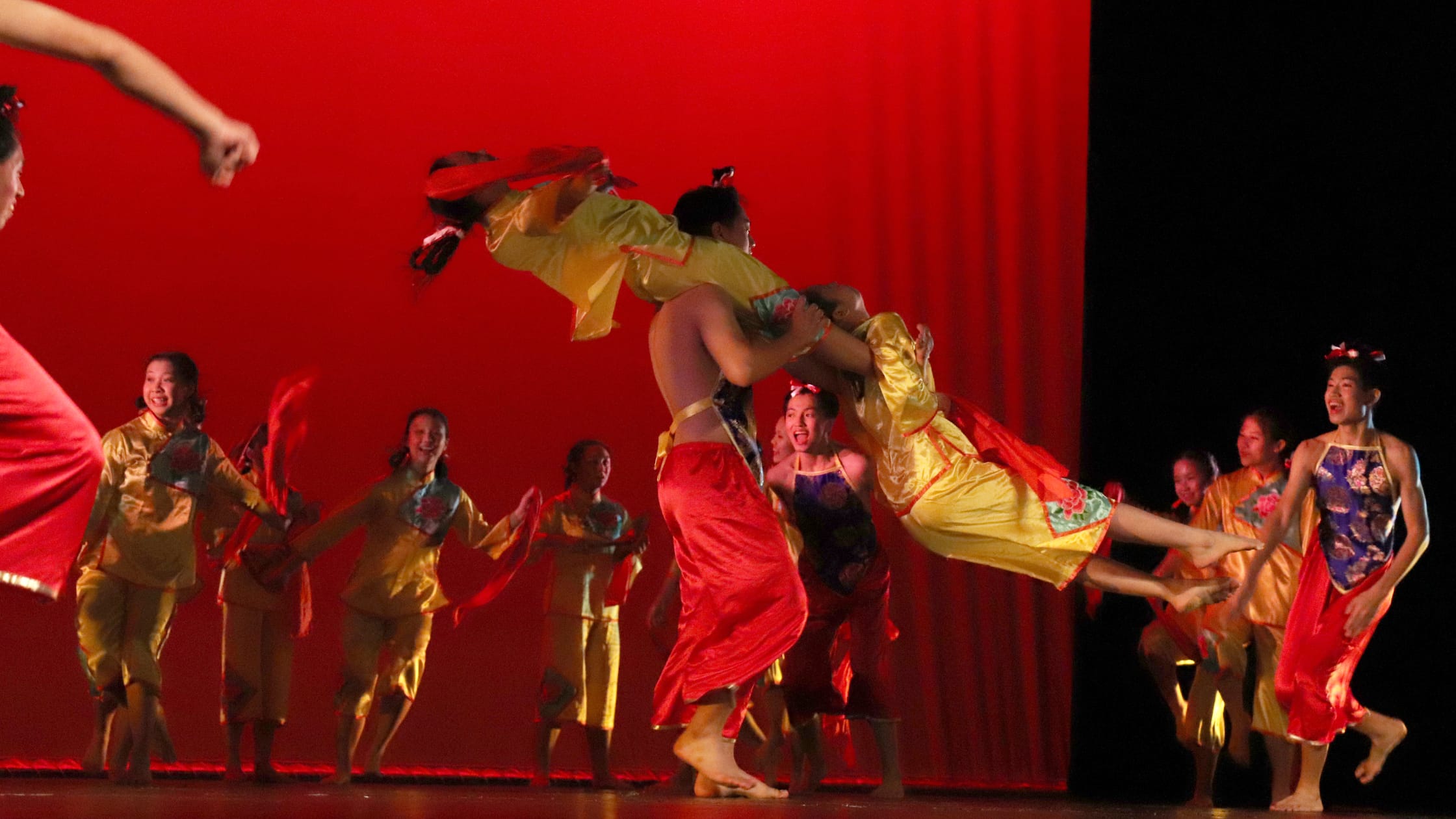 Performers in yellow and red traditional Chinese costumes dance on stage, one being lifted, against a red backdrop.