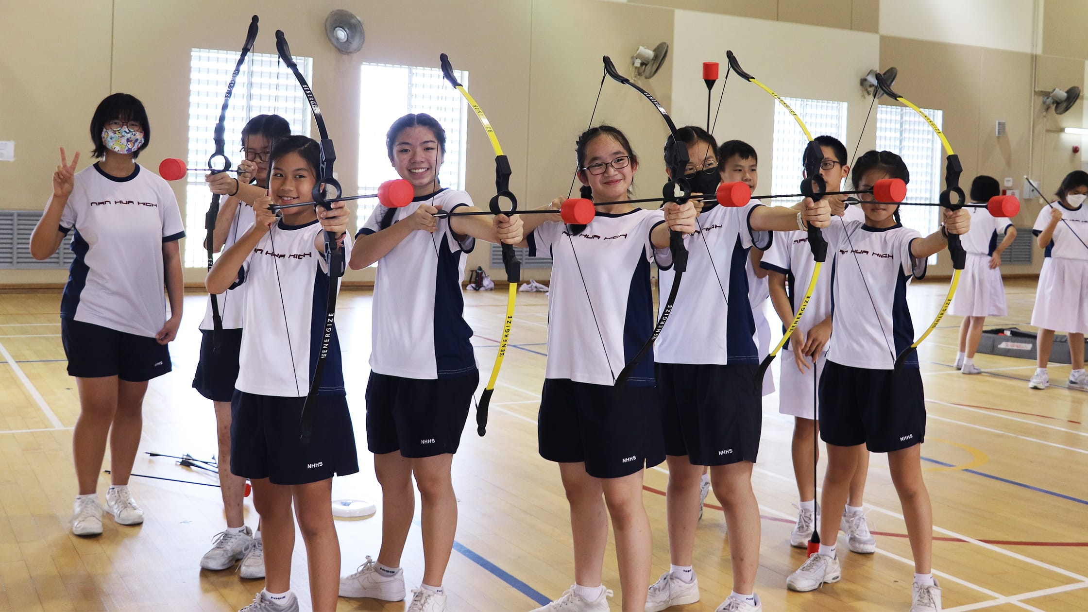 Students in school uniforms aim yellow and black bows with red-tipped arrows in an indoor gymnasium.