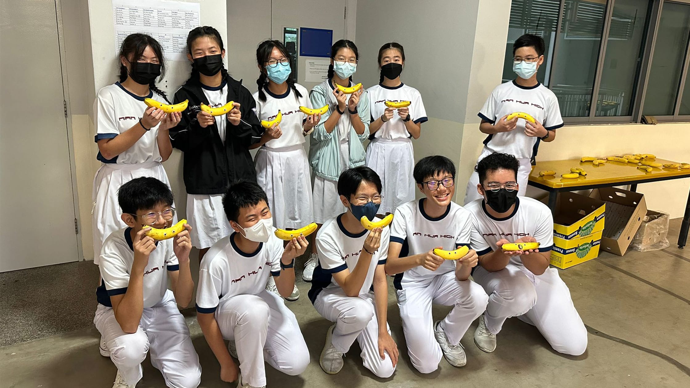Group of students in white uniforms and face masks posing together, each holding a yellow banana toward the camera.