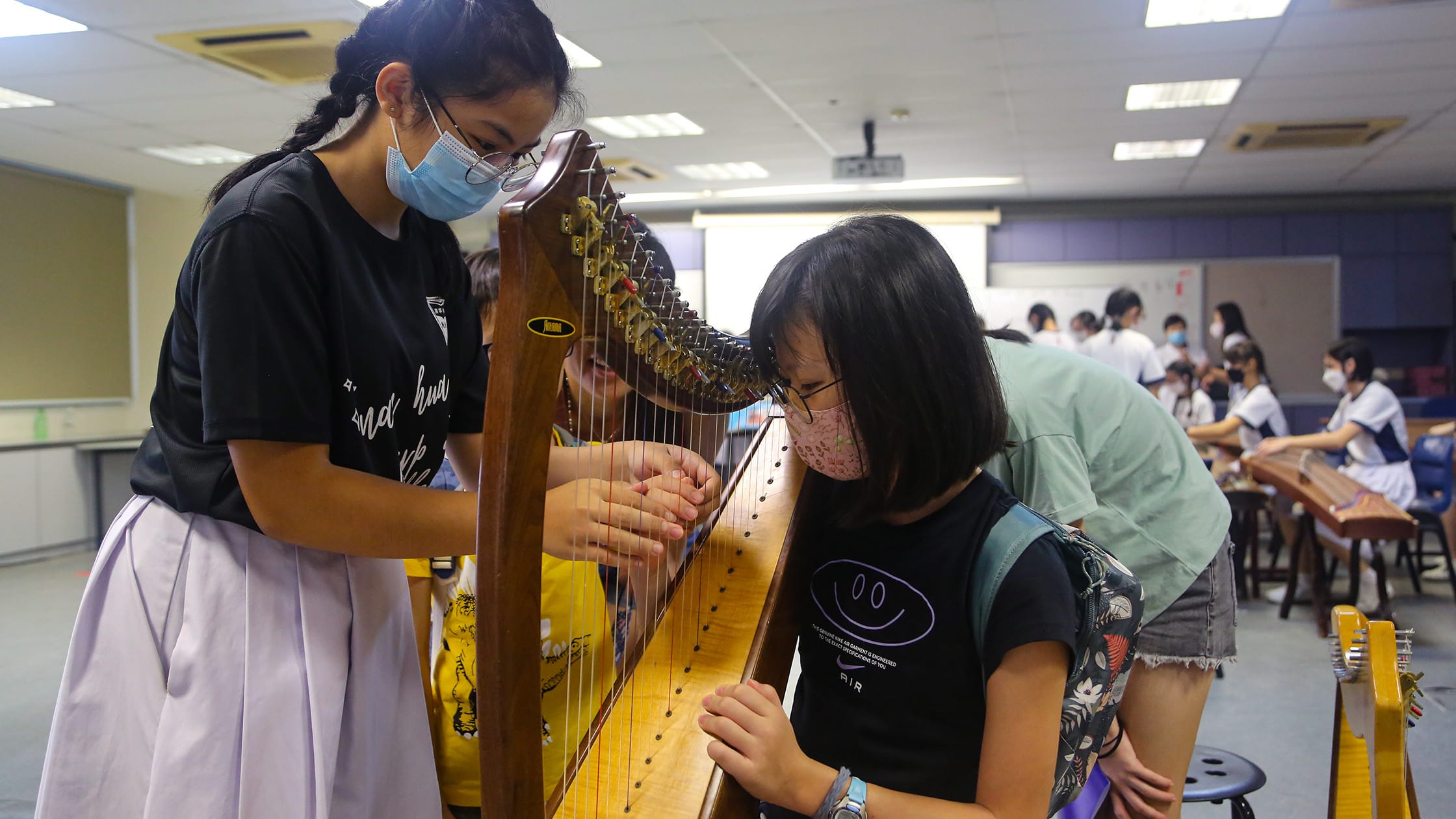 Two masked girls examine a wooden harp closely in a classroom where other students play string instruments.