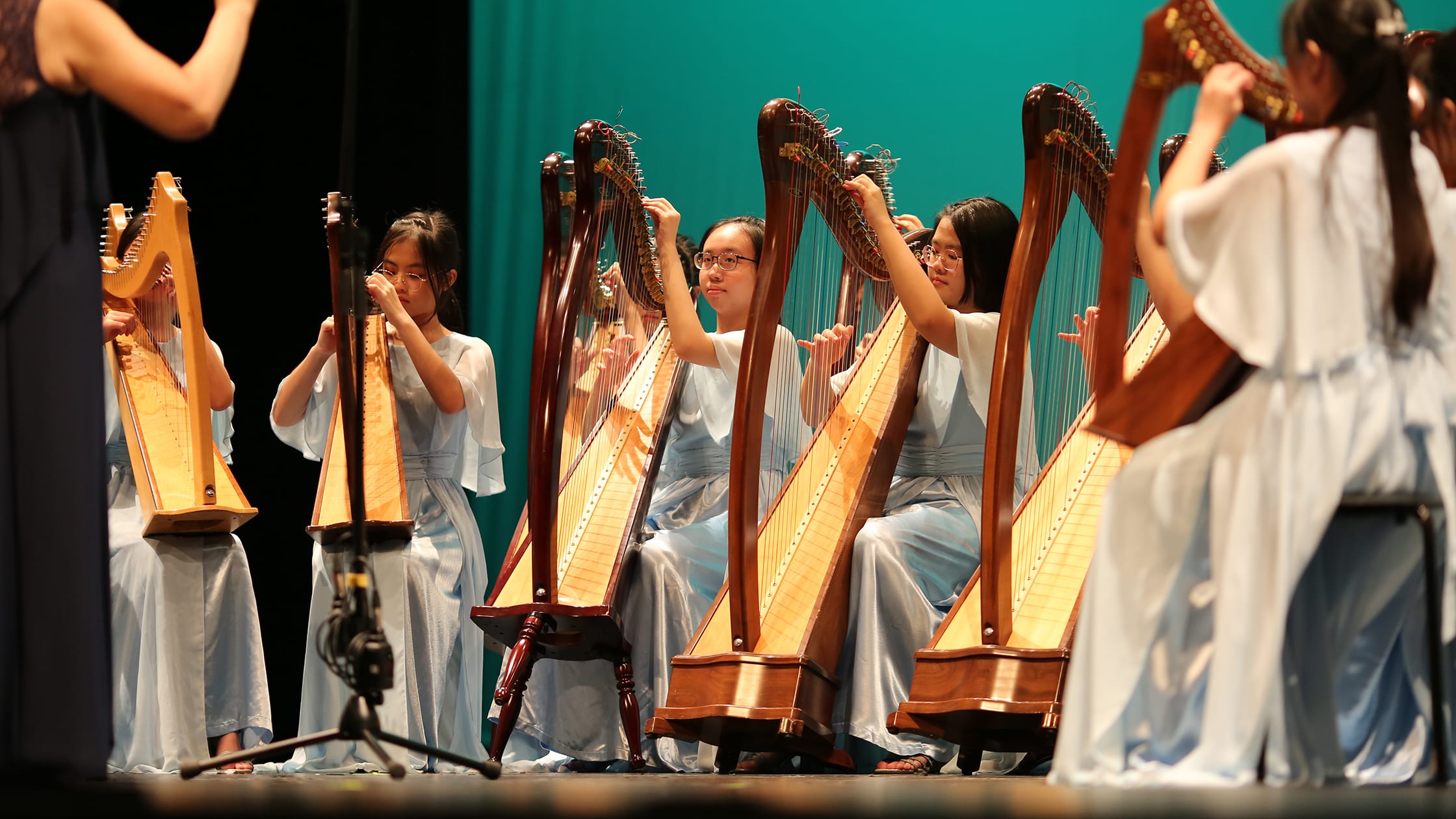 Several young girls in white dresses sit on stage playing harps before a teal backdrop.