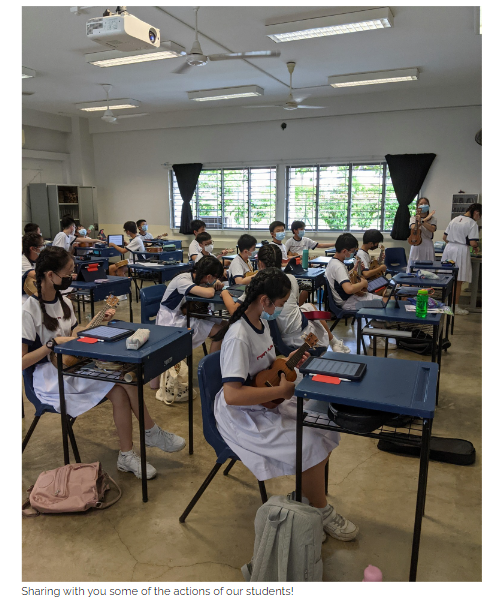 Students in white uniforms and masks sit at spaced desks in a classroom, each holding a ukulele.