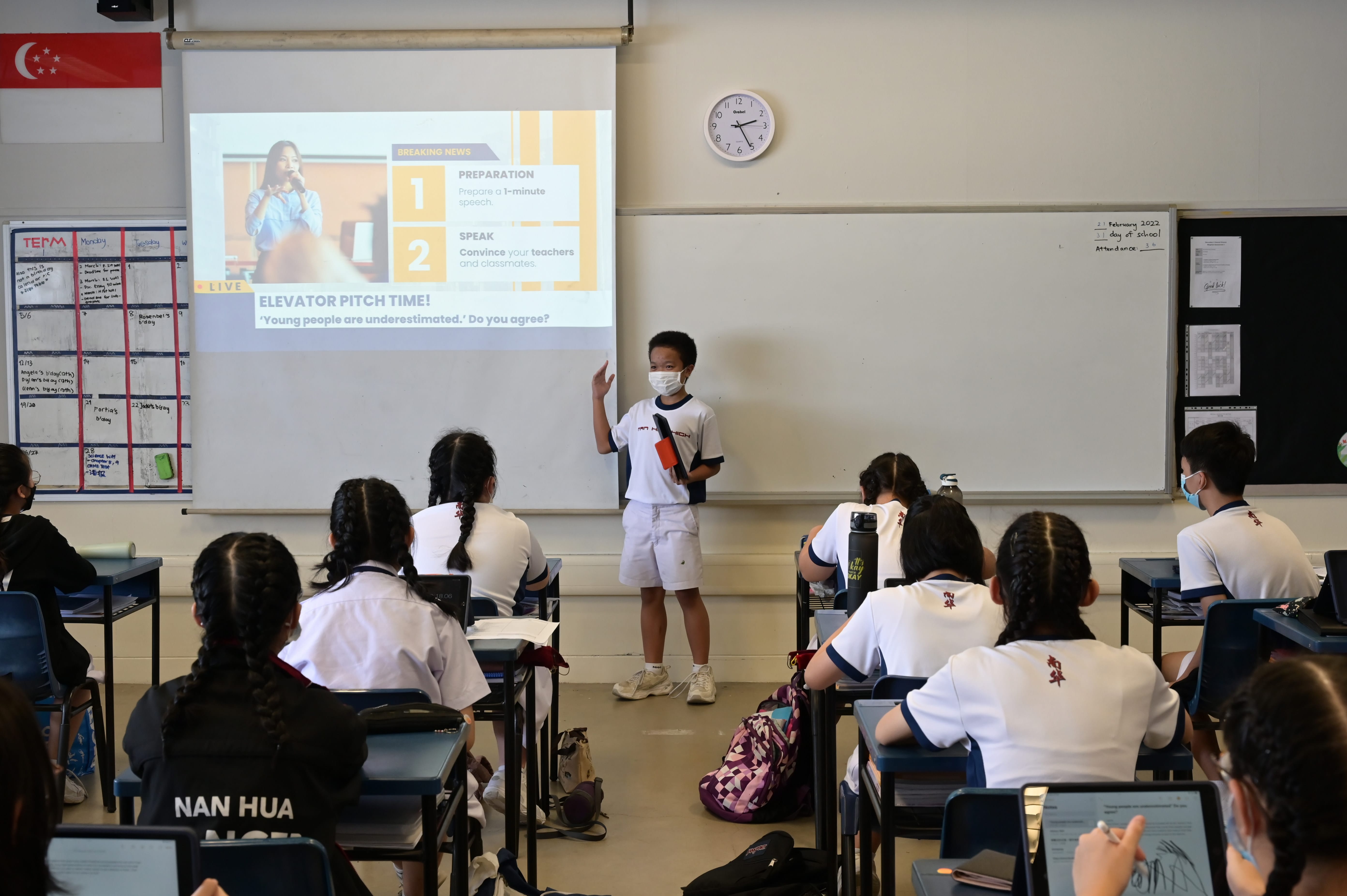 A masked student holds a microphone presenting an elevator pitch to classmates in a school classroom with a projector screen.
