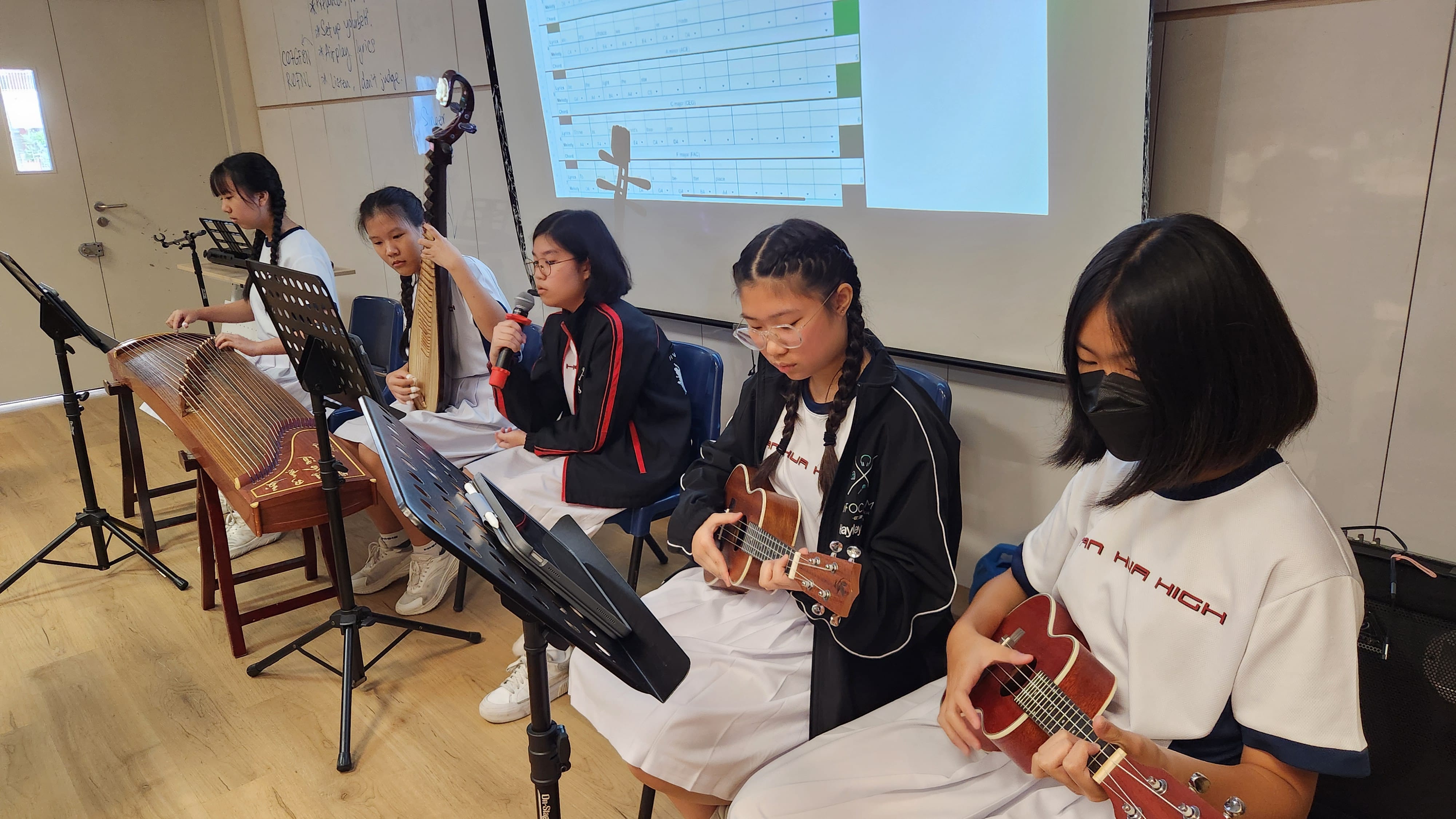Five school girls perform in a classroom, playing guzheng, erhu, ukuleles, and singing into a microphone.