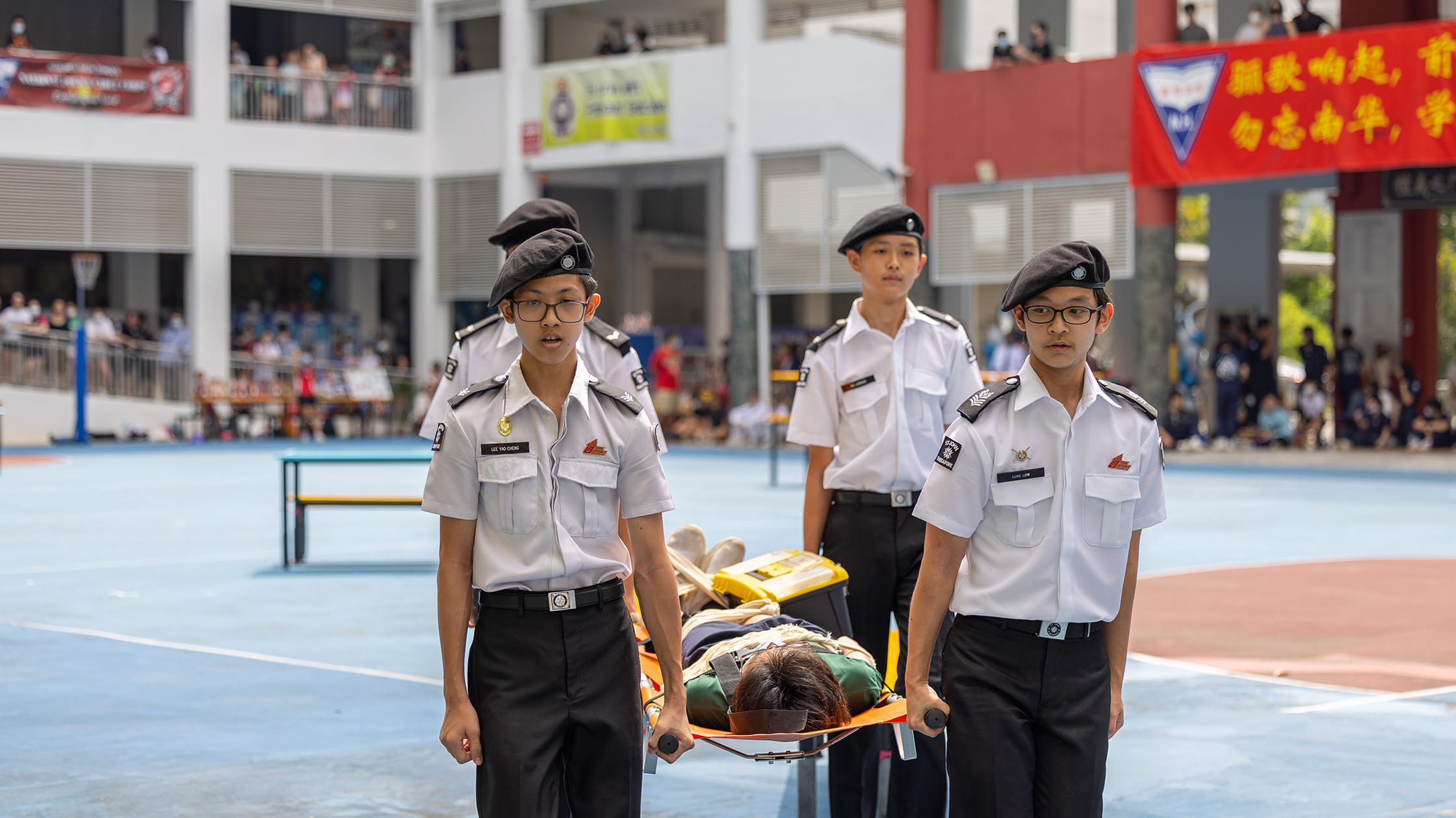Three uniformed cadets carry a person on a stretcher across a school courtyard during a public demonstration.