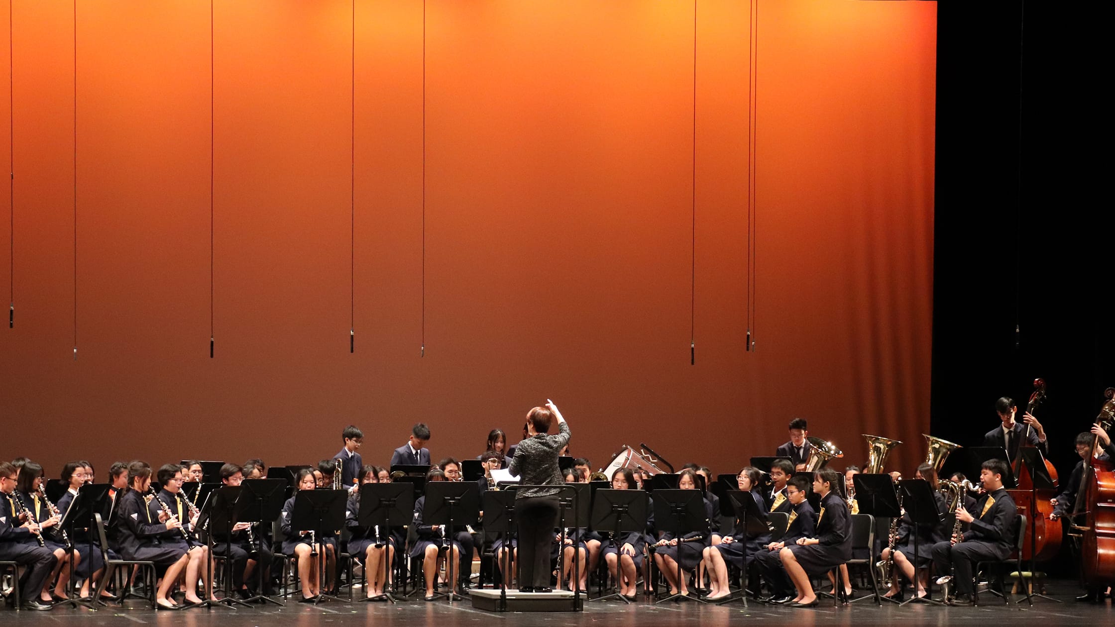 A conductor leads a large youth wind band on stage before an orange backdrop, all wearing dark uniforms.