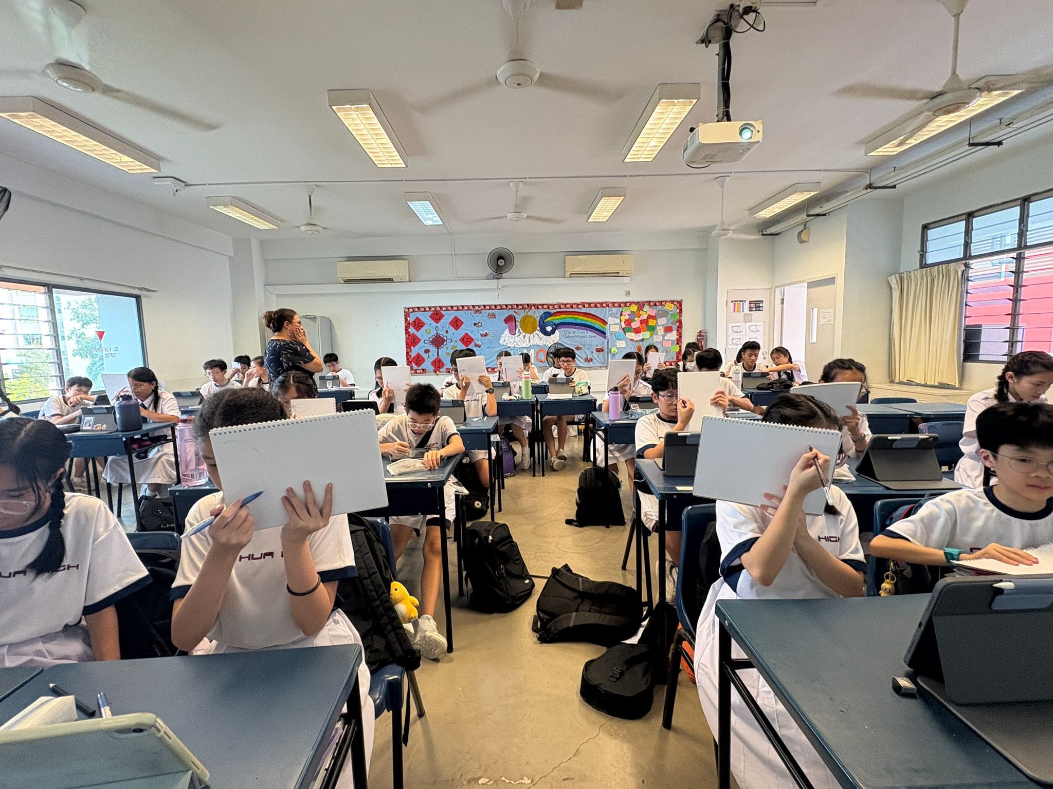 Students in white uniforms sit at desks holding up sketch pads in a decorated classroom with a teacher standing at the back.