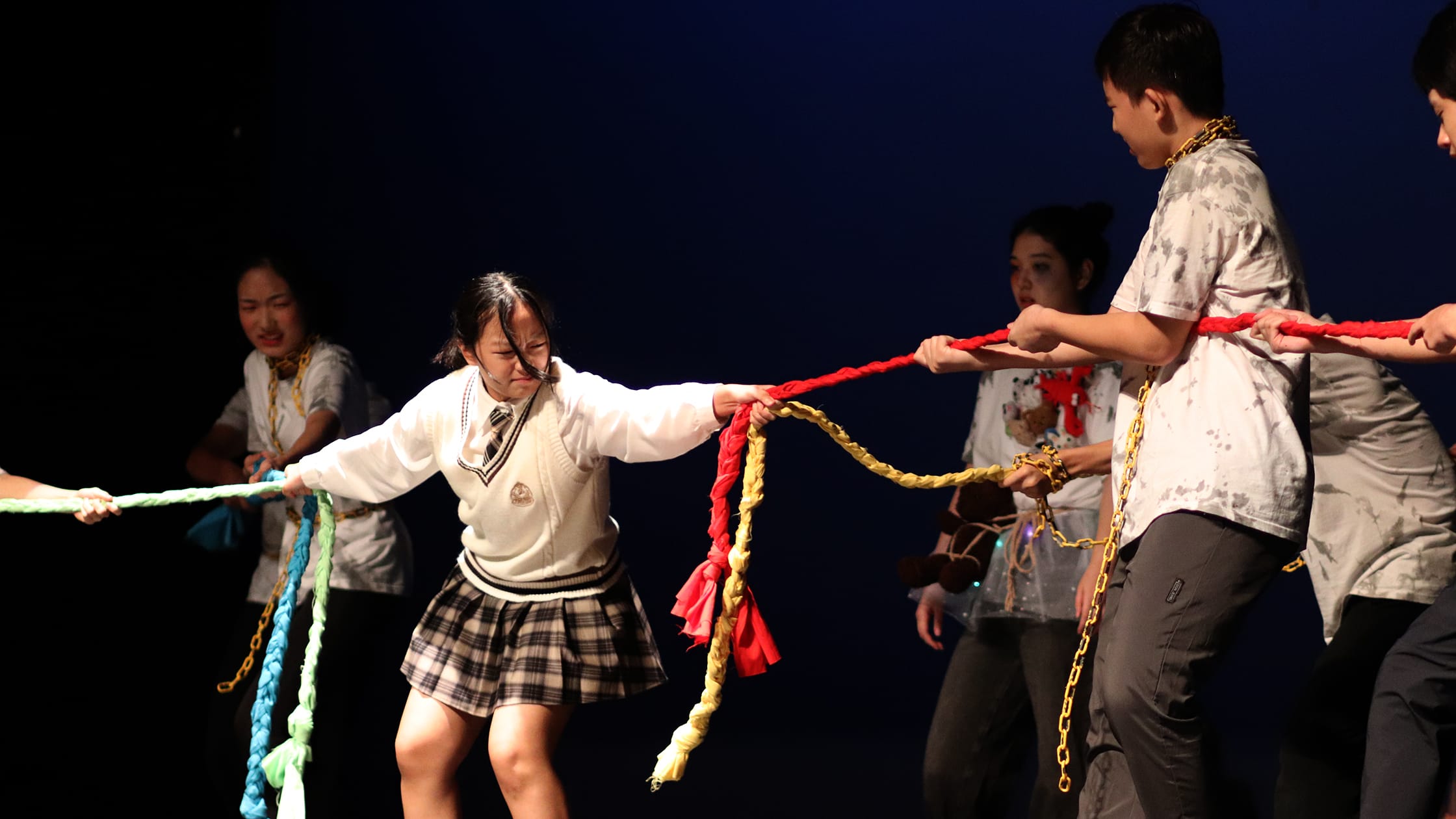 Students perform on a dark stage, pulling colorful braided ropes and chains in a theatrical scene.