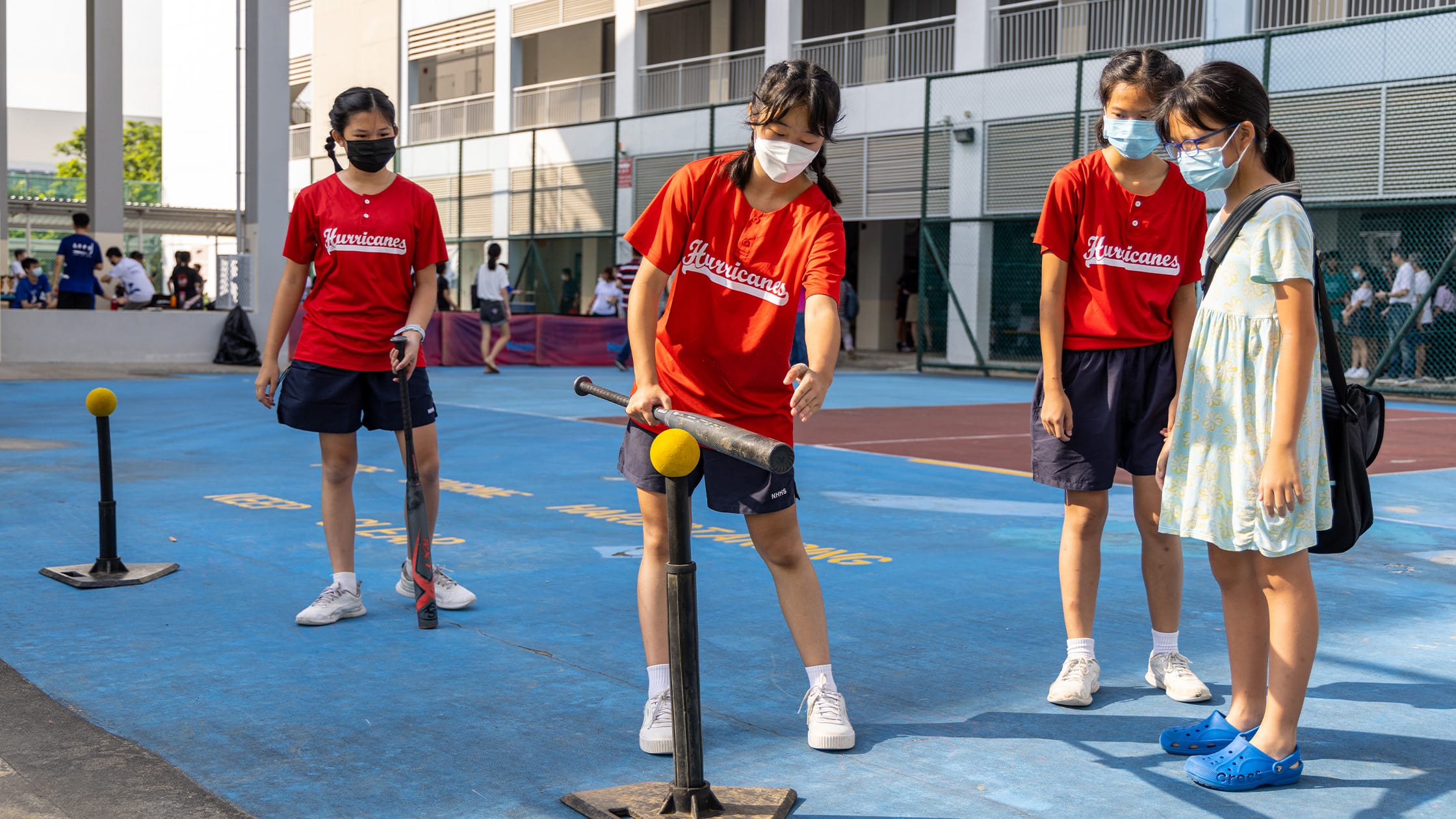 Four masked girls on a blue court, one swinging a bat at a yellow ball on a tee, wearing Hurricanes jerseys.