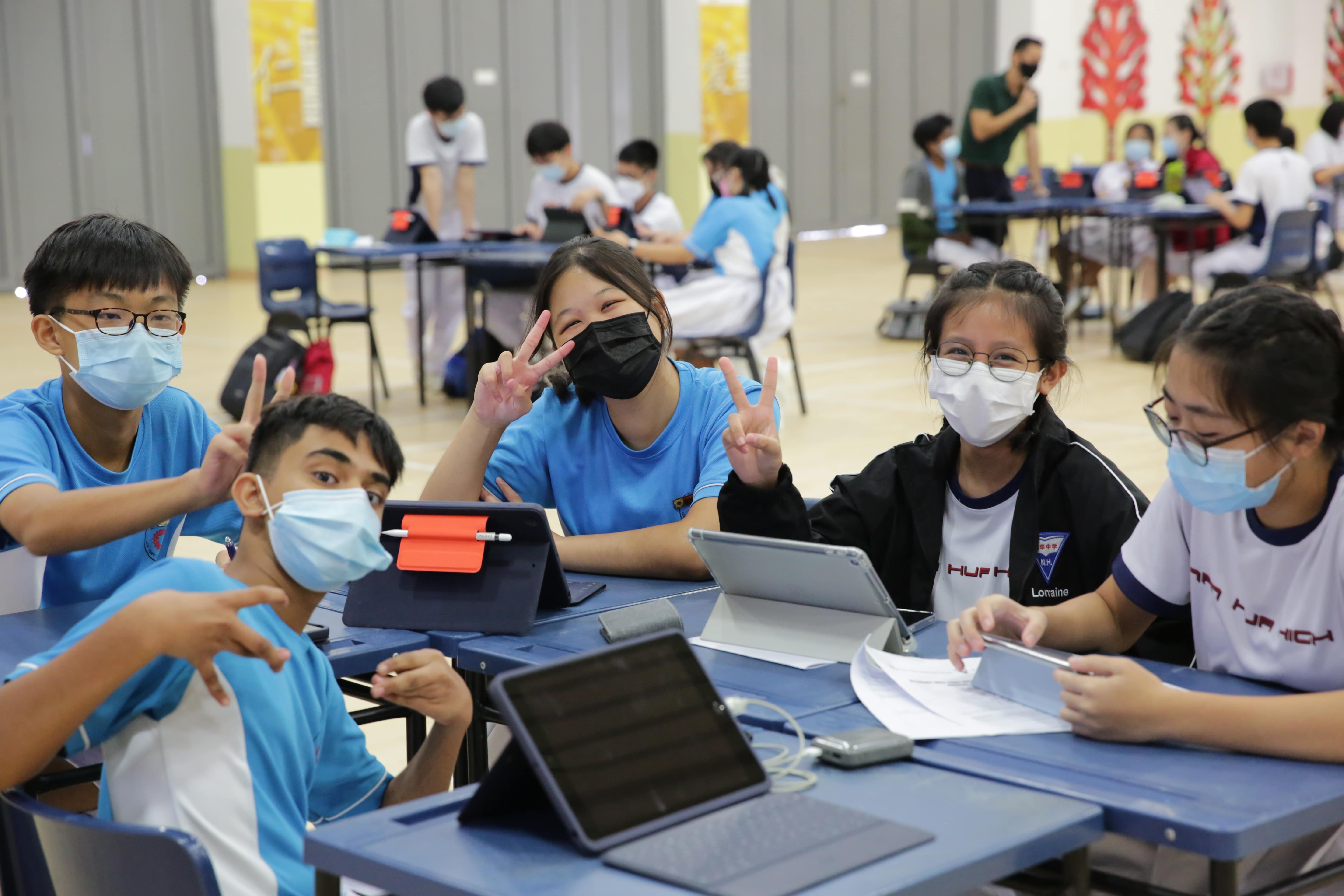 Students wearing face masks sit at blue desks with tablets, smiling and making peace signs toward the camera.