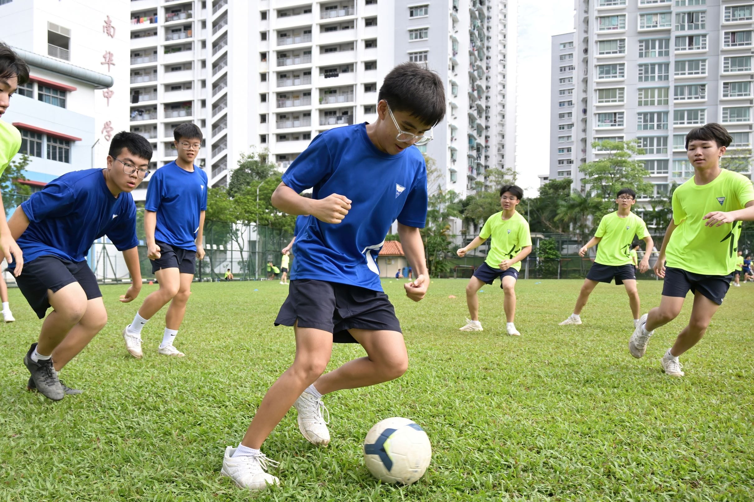 Teen boys in blue and green jerseys play soccer on a grass field with tall apartment buildings in the background.
