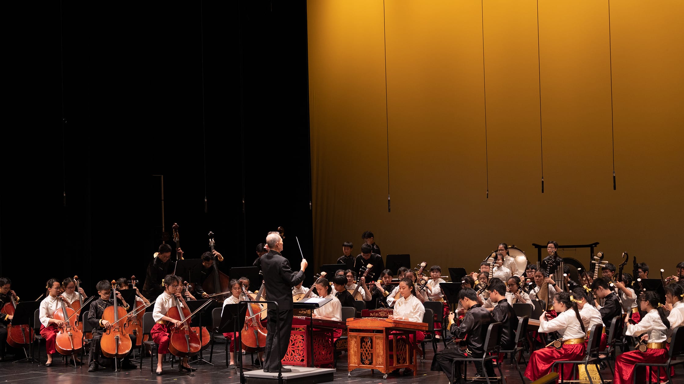 A conductor leads a large youth Chinese orchestra on a warmly lit stage with traditional and string instruments.