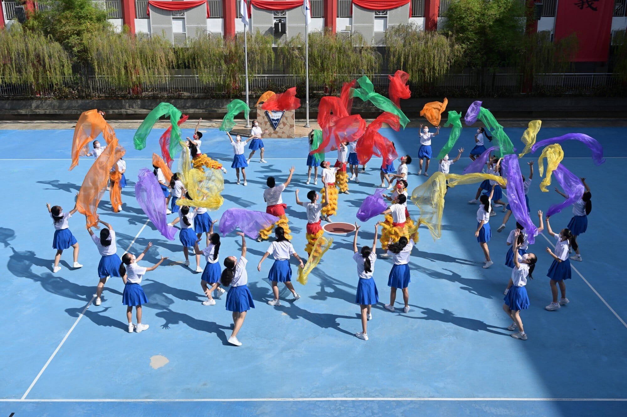 Students in white shirts and blue skirts wave colorful scarves on a blue outdoor court during a performance.