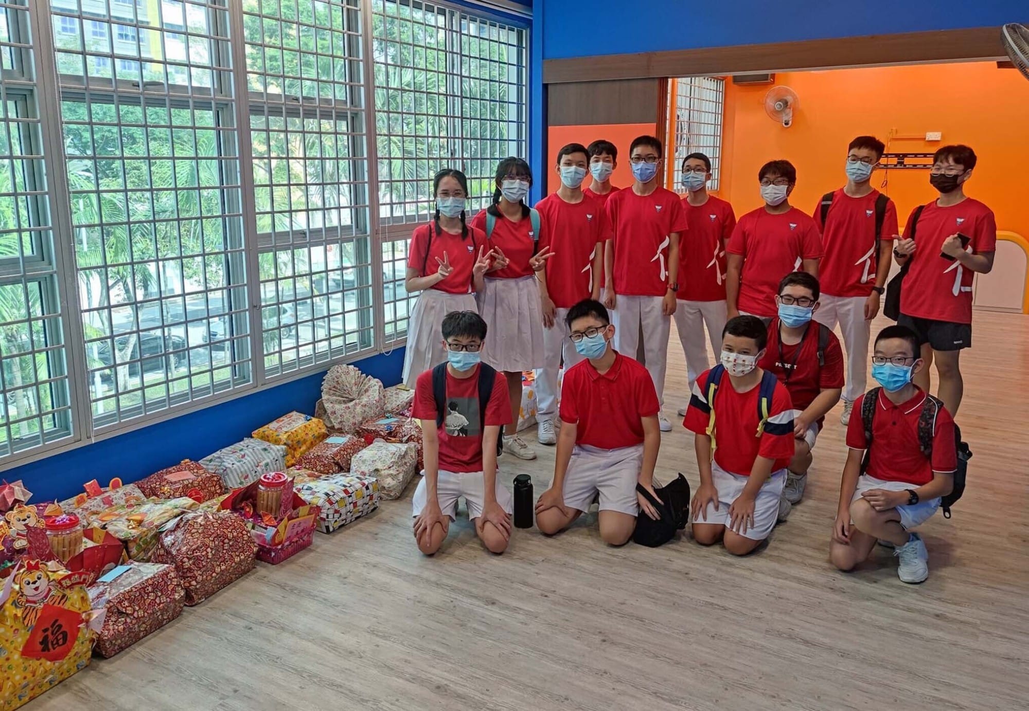 A group of students in red uniforms wearing masks pose indoors beside wrapped gift boxes and festive bags.