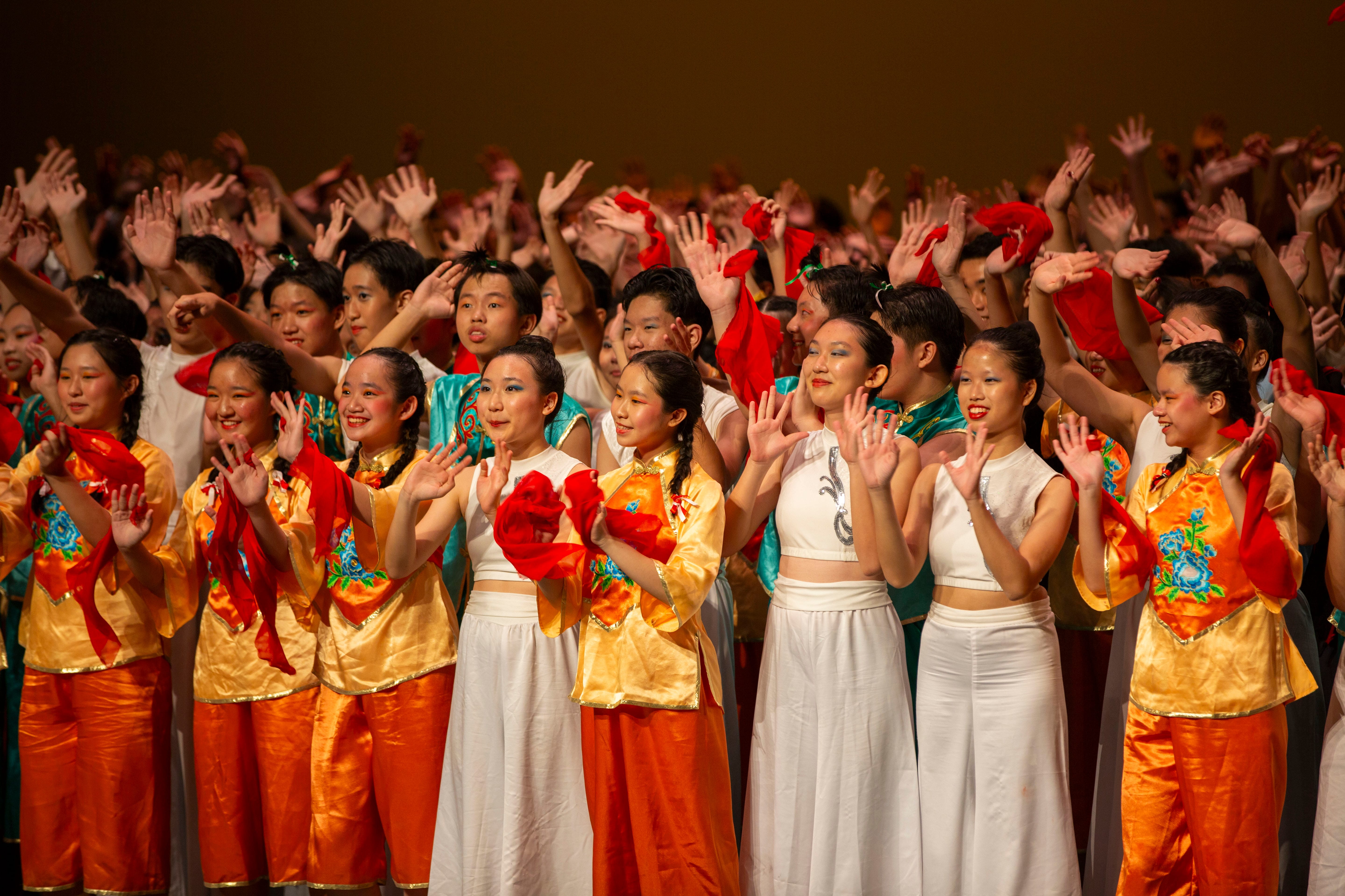 Large group of performers in gold, orange, and white traditional Chinese costumes wave hands and smile on stage.