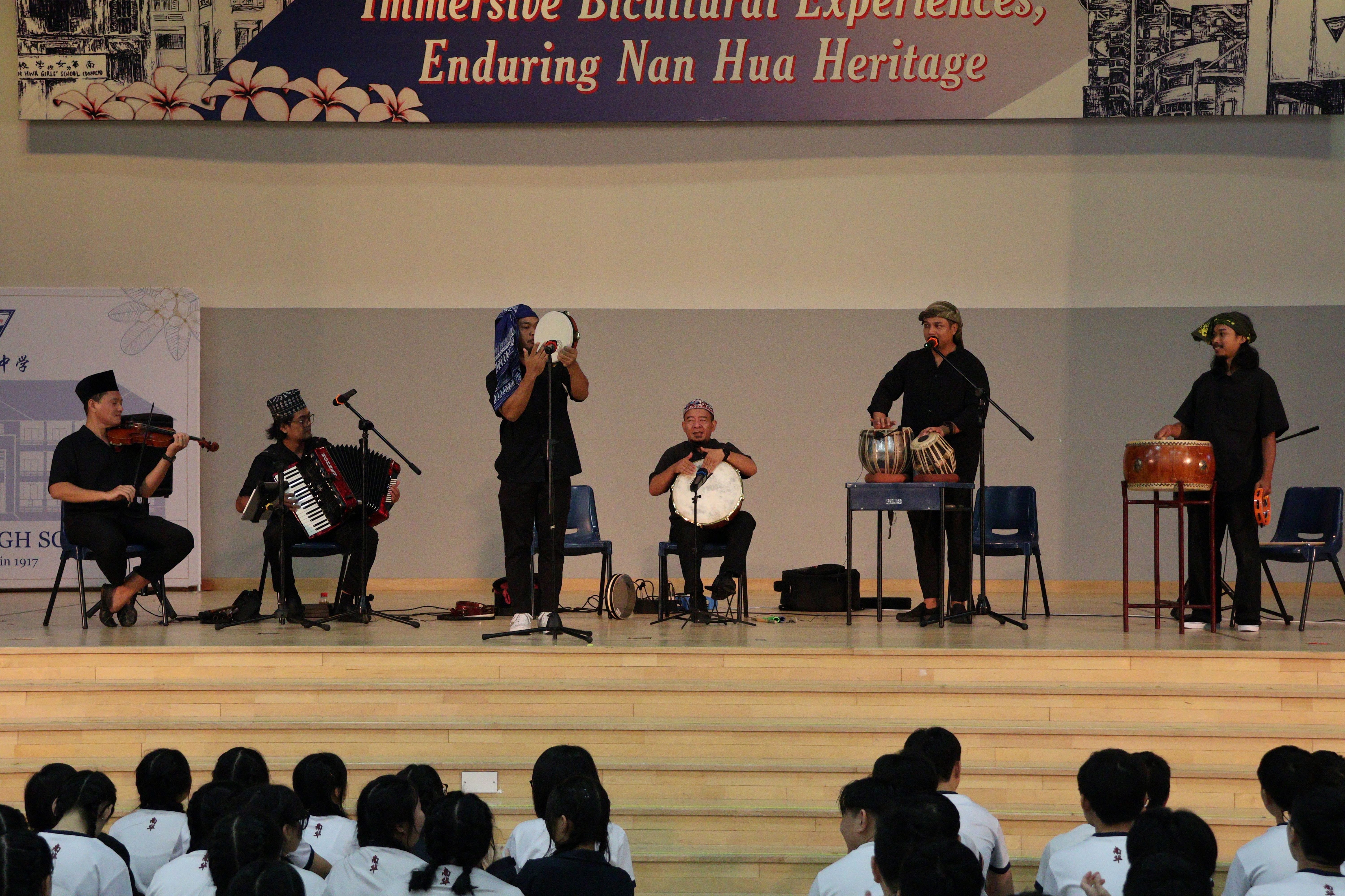 Six musicians in black performing with drums, violin, and accordion on stage at Nan Hua Heritage school event.