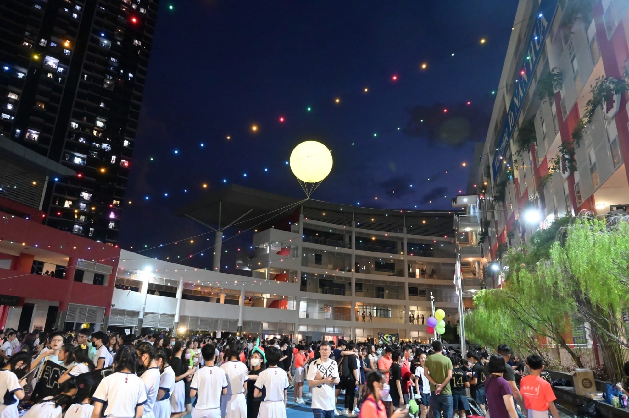 Large crowd gathers at night in a schoolyard with colorful lights and a glowing moon balloon floating above.