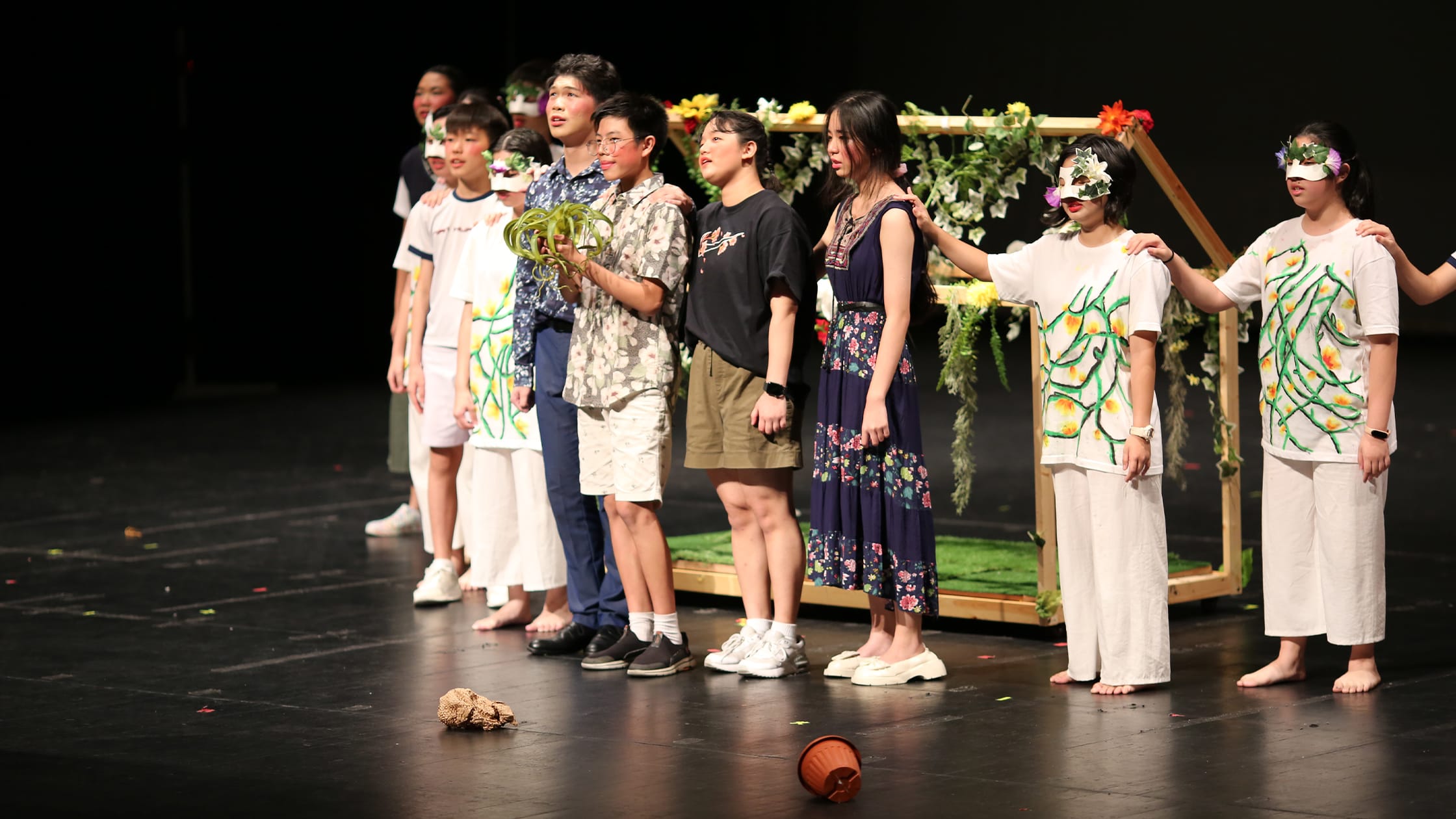 Young performers in floral costumes and leaf masks stand together on a dark stage with a flower-decorated wooden arch prop.