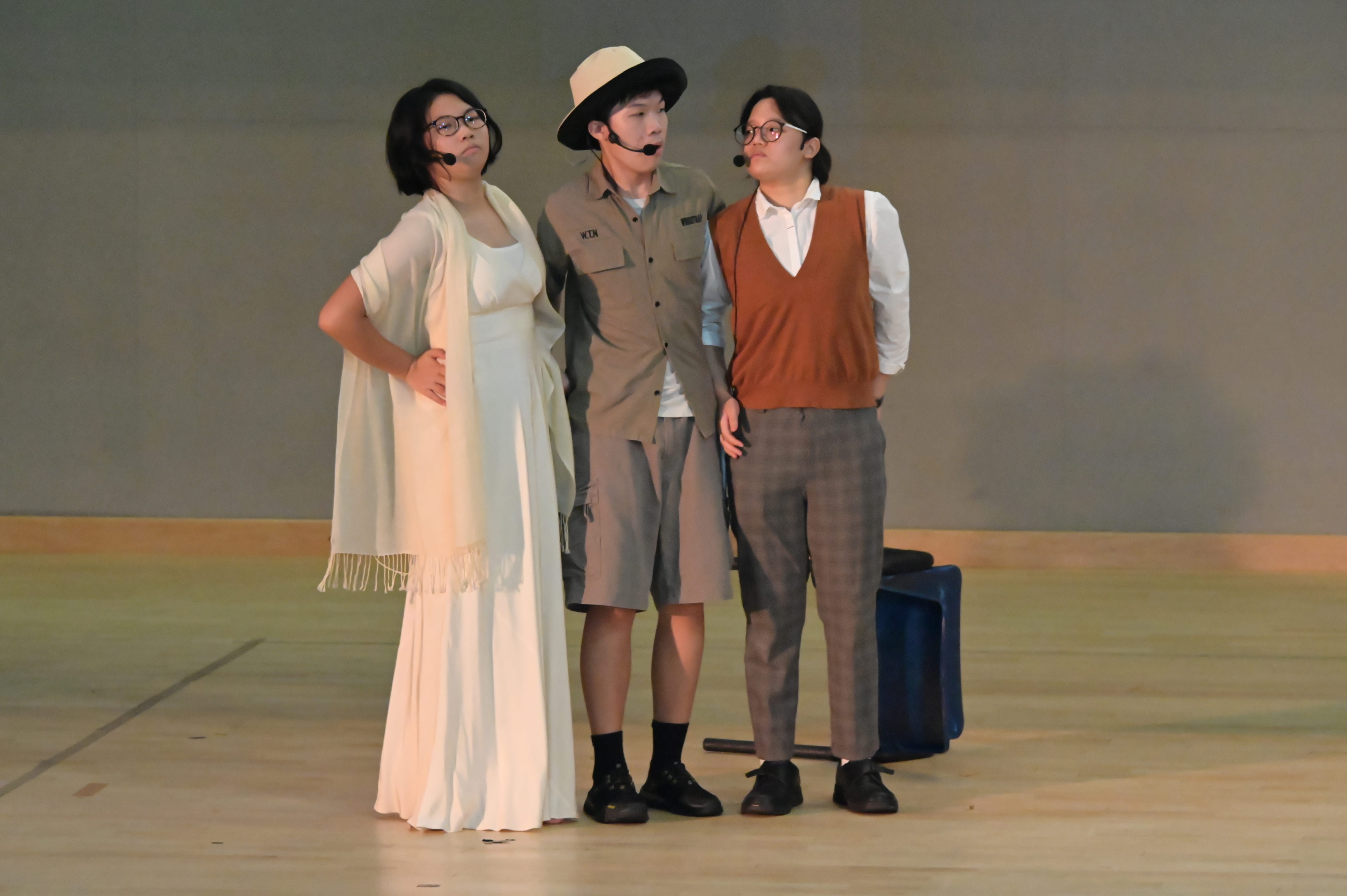 Three performers on stage wearing microphones, costumes including a white dress, safari outfit with hat, and brown vest.