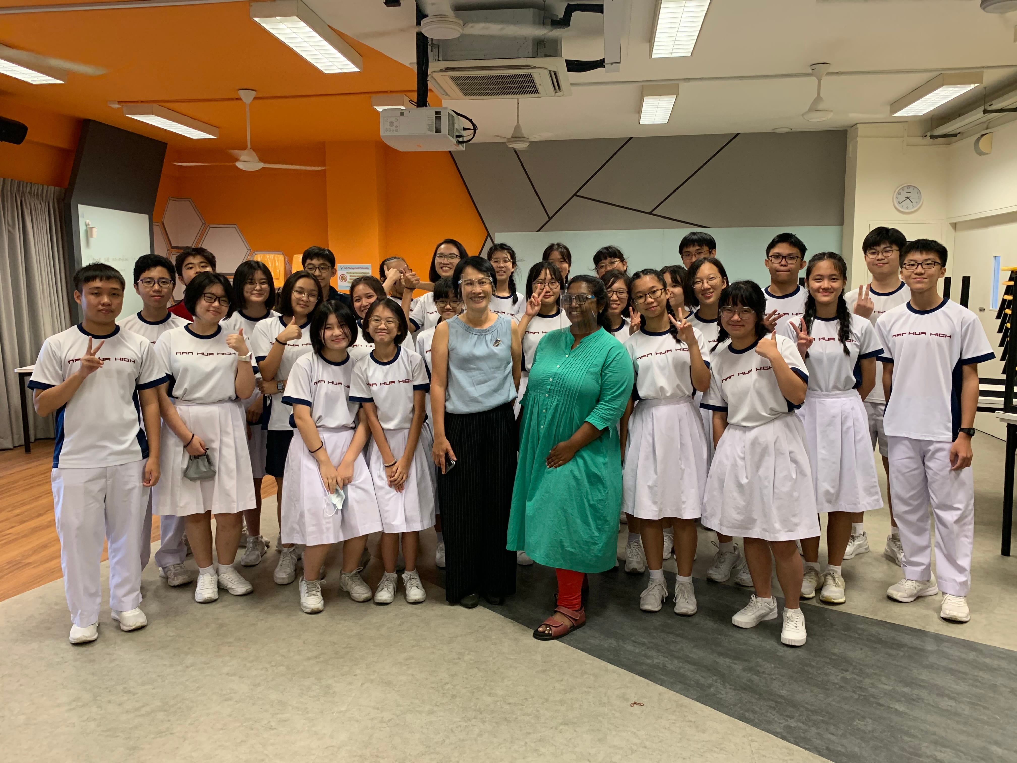 Two adults pose with a large group of students in white Nan Hua High School uniforms inside a classroom.