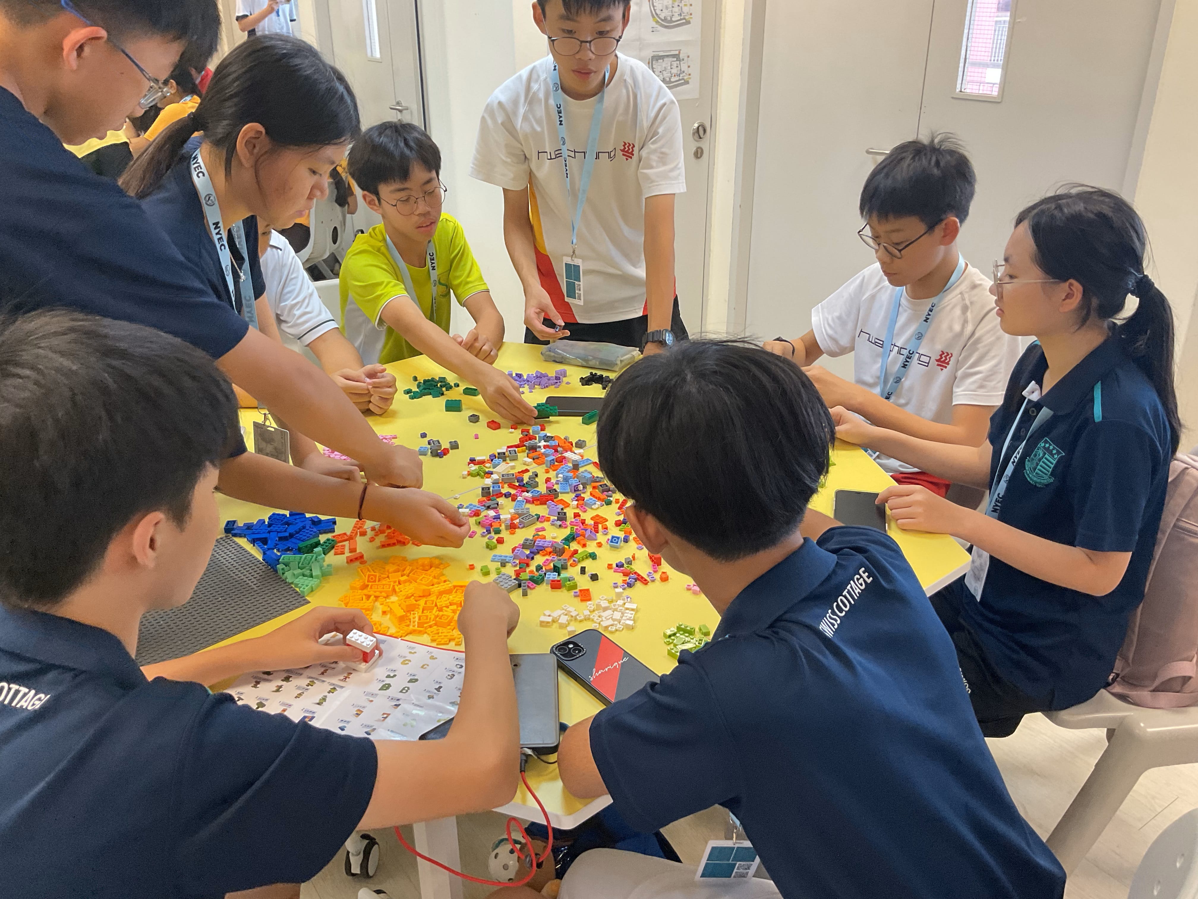 Group of students around a yellow table sorting colorful plastic bricks, with instruction sheet and phones nearby.