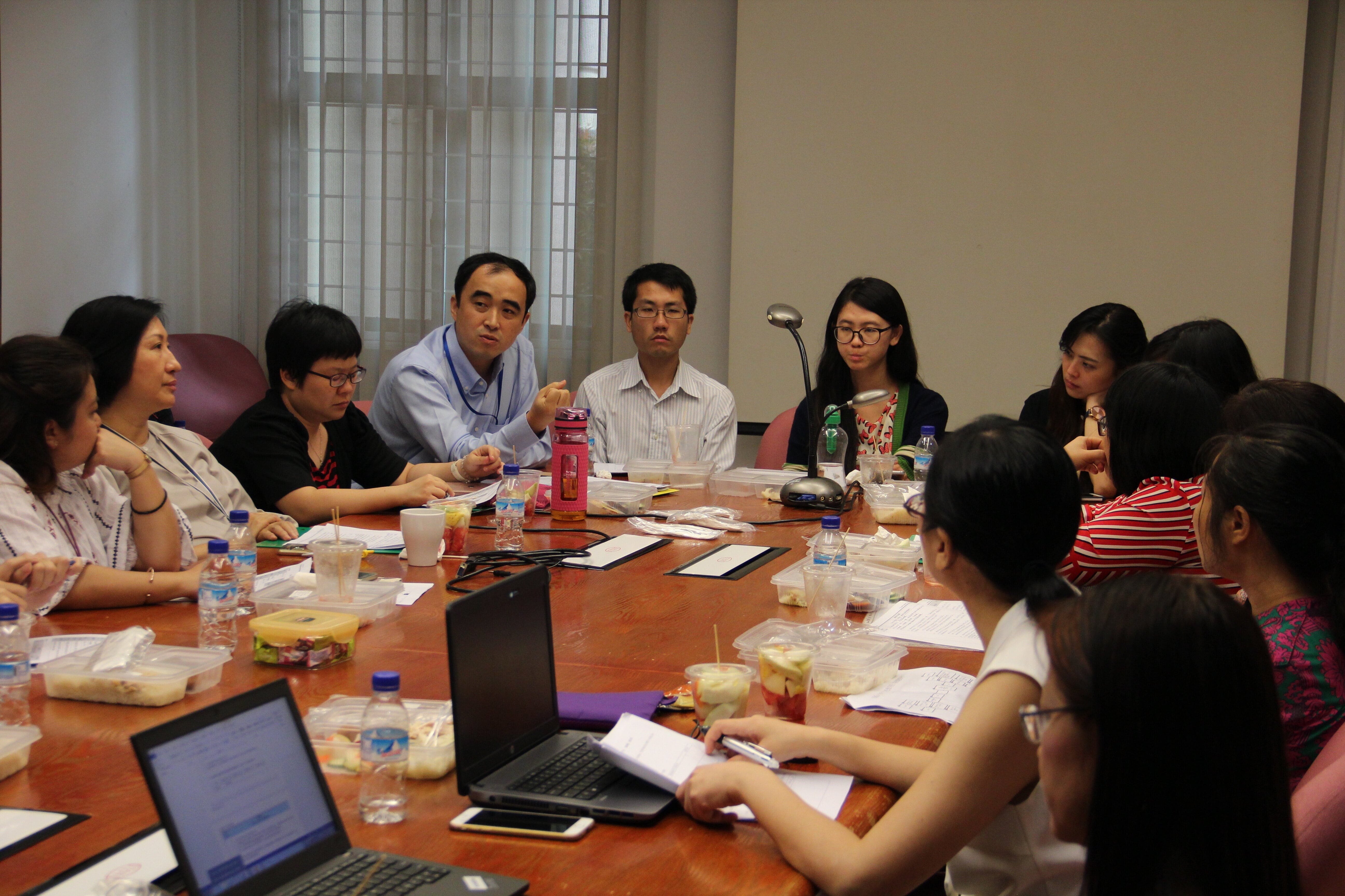 A group of people sit around a large wooden conference table with laptops, papers, food, and drinks during a meeting.