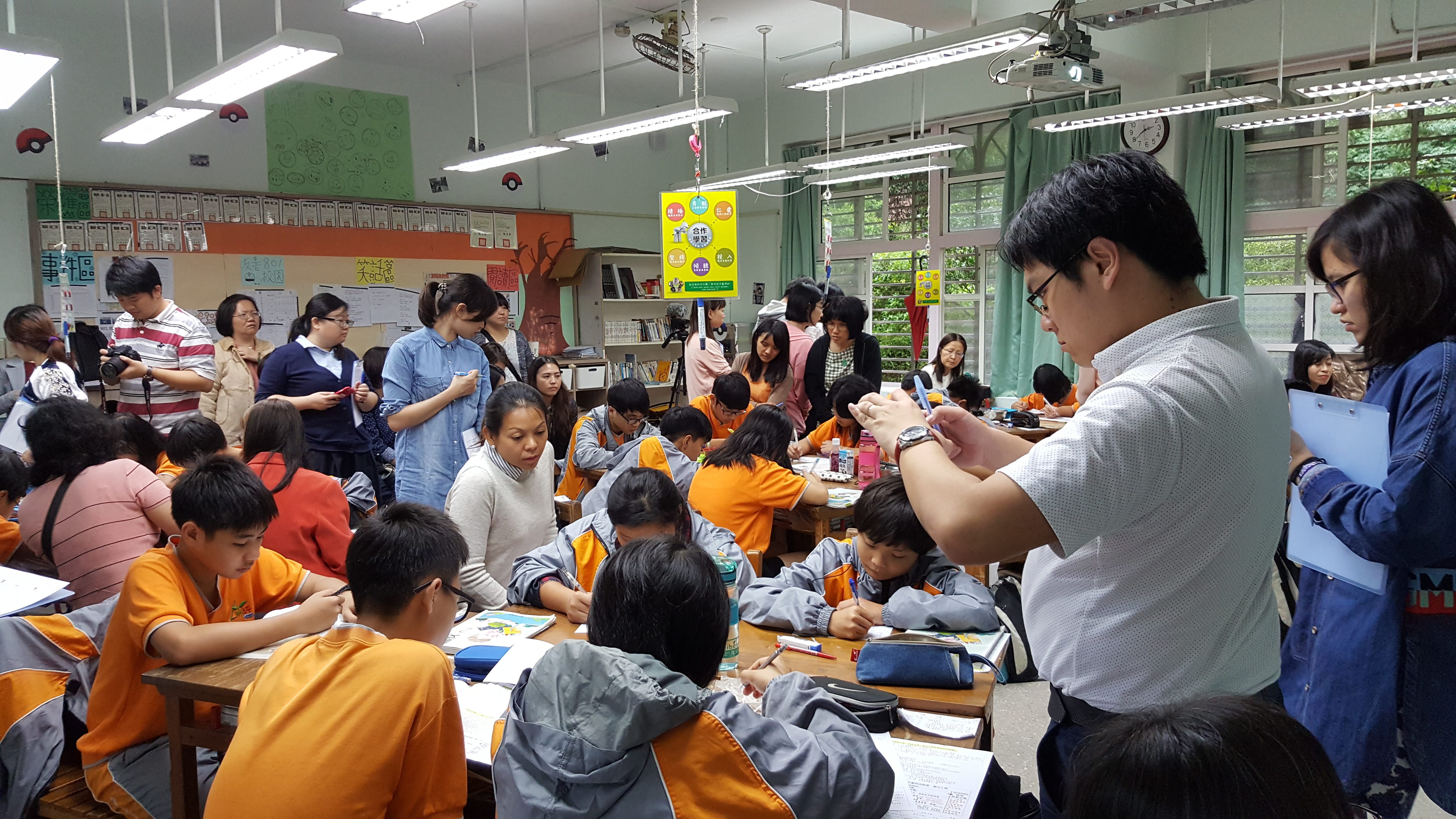 Busy classroom with students in orange uniforms working at tables while adults observe and take photos