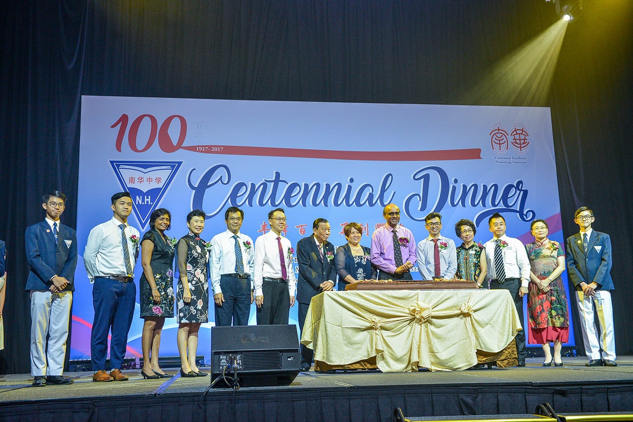 A group of people stand on stage at N.H. school's 100th Centennial Dinner event with a cake on a table.