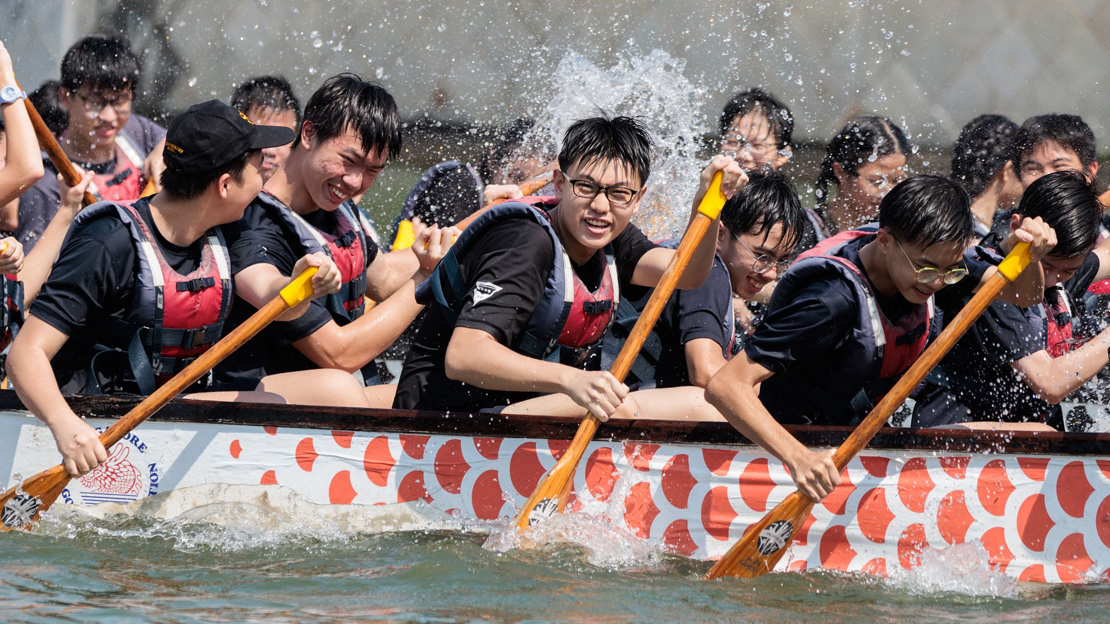 A team of young paddlers in life vests rowing a dragon boat with wooden oars, water splashing around them.