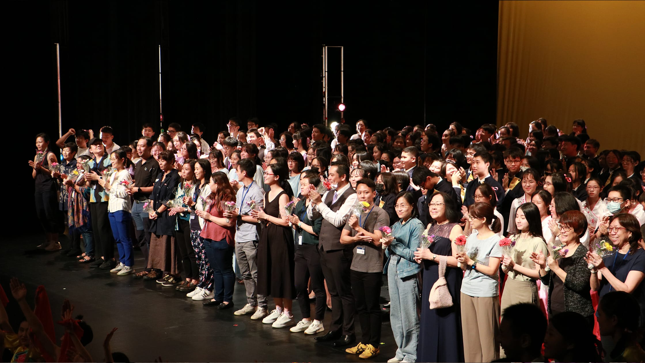 A large group of people standing on a dark stage, clapping and holding flowers during a curtain call.