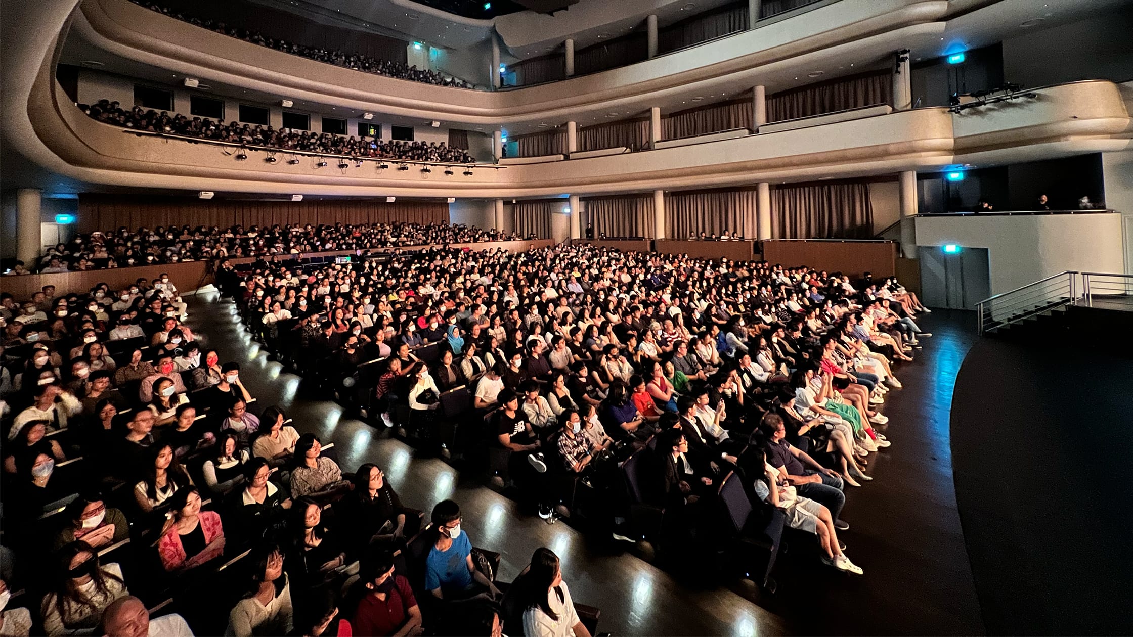 A packed multi-level theater auditorium filled with hundreds of seated audience members facing a stage.