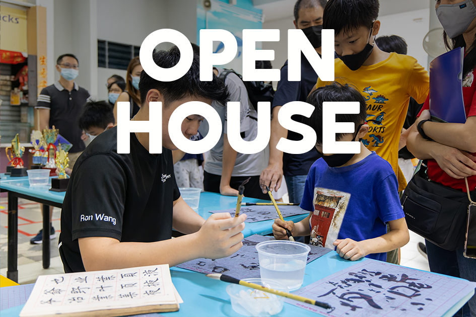 Children practice Chinese calligraphy with brushes at a school open house event, surrounded by masked adults watching.