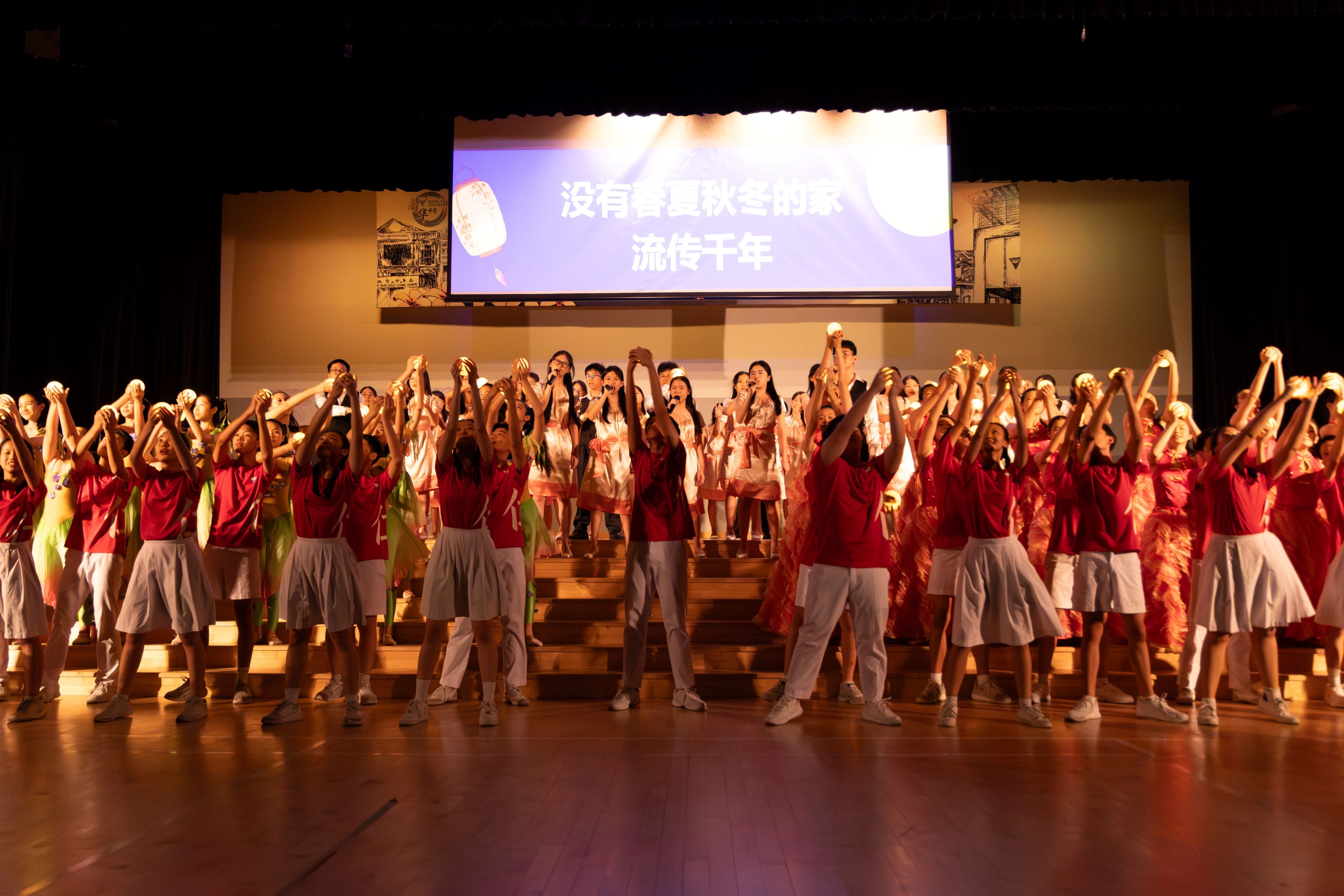 Students in red shirts and white bottoms raise glowing objects on stage before a screen with Chinese text.