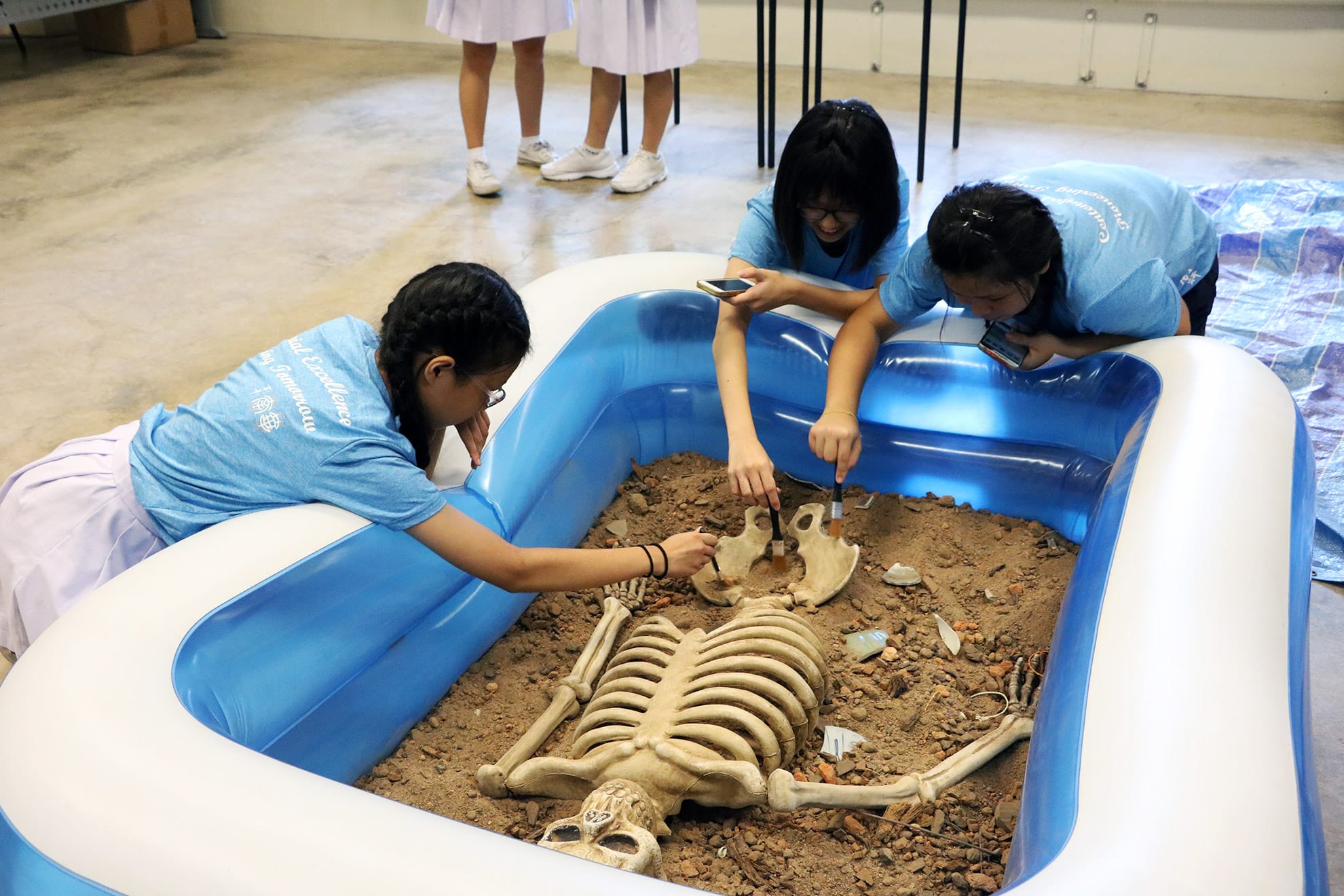 Three students lean over an inflatable pool, brushing dirt from a human skeleton in a classroom.