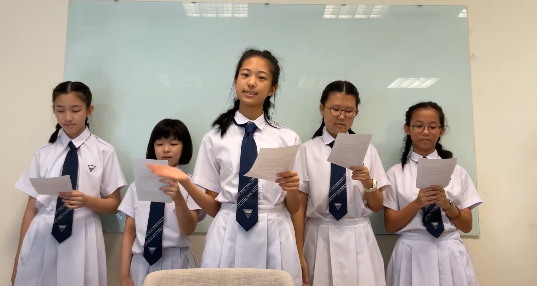 Five girls in white school uniforms with navy ties stand together, each holding papers as if reading aloud.