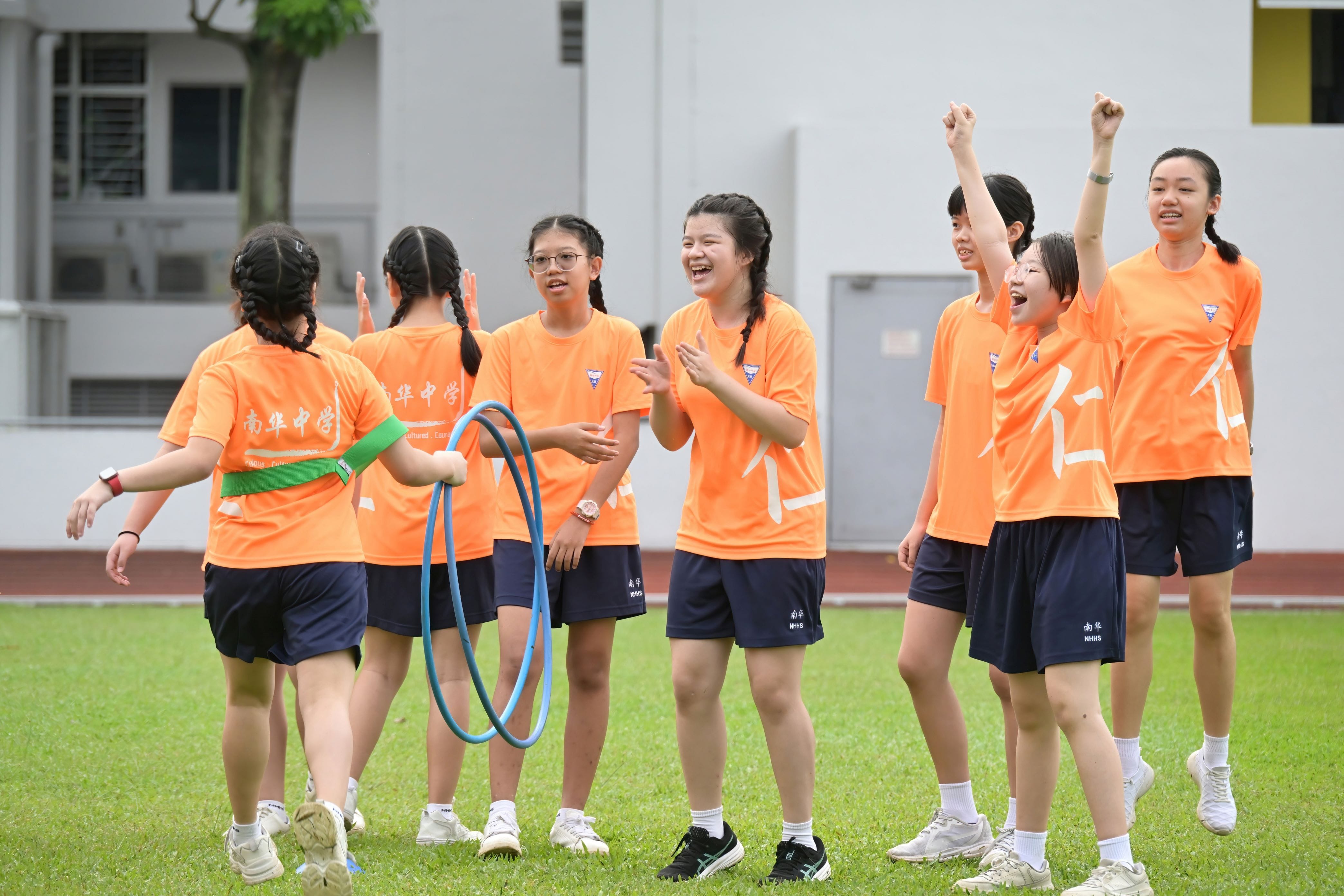 Seven girls in orange school uniforms celebrate on a grass field, some raising fists and clapping, holding blue hula hoops.