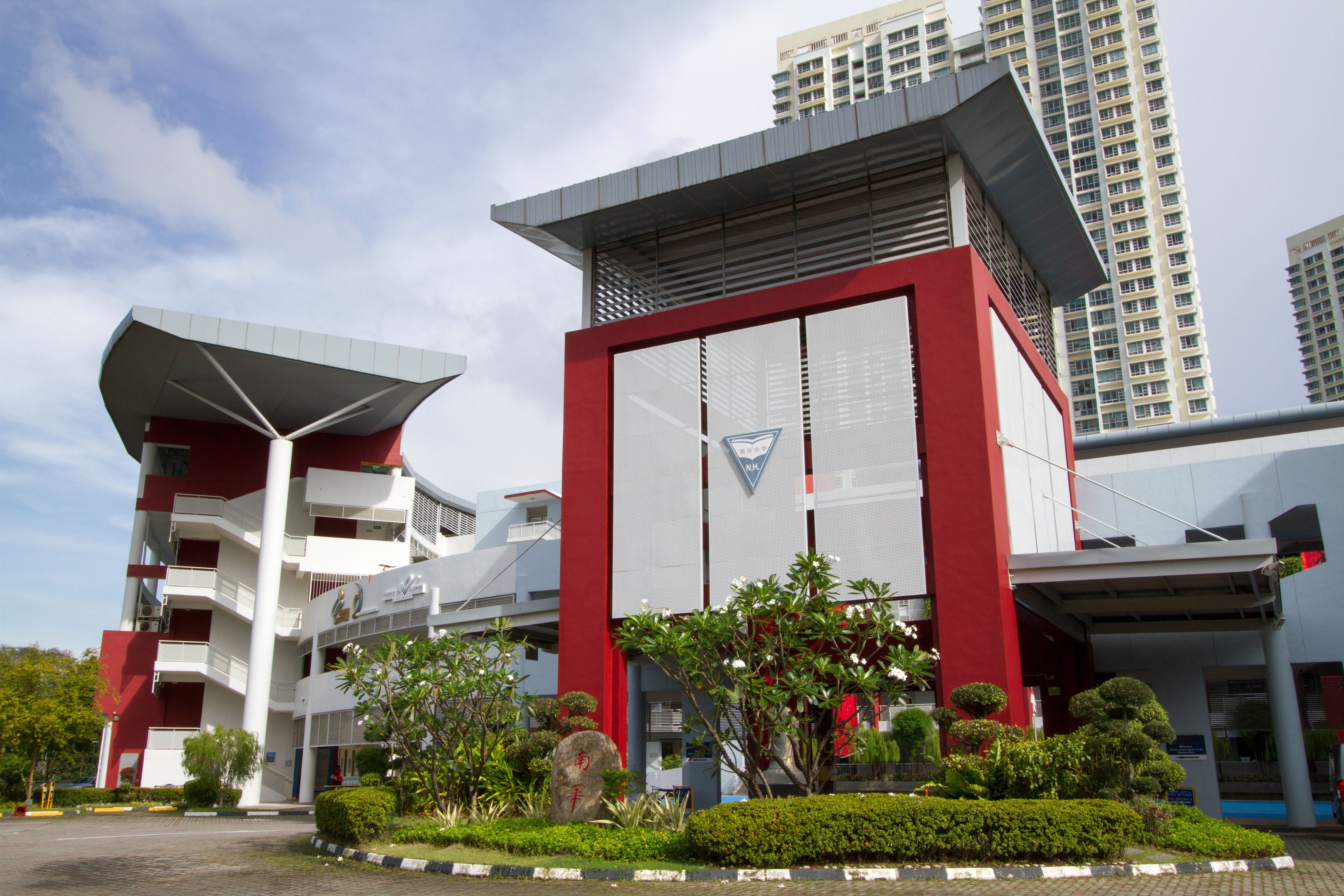 Modern red and white school building with a large entrance tower, landscaped shrubs, and tall residential buildings behind.