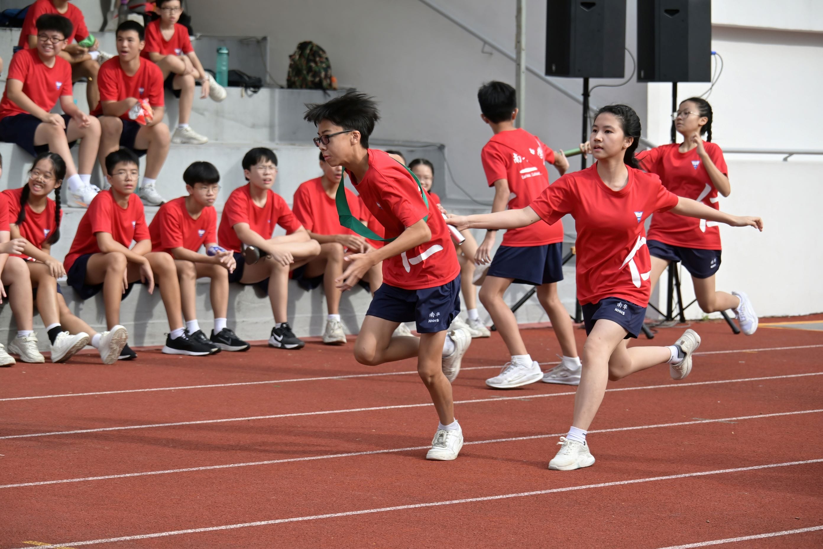 Students in red shirts pass a baton during a relay race on a track, with teammates watching from bleachers.