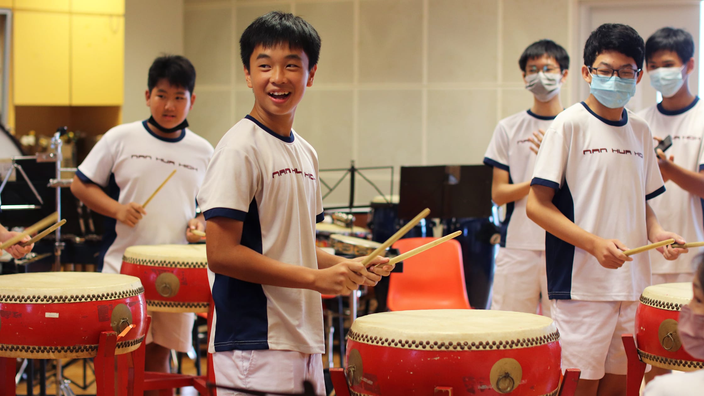Several young students in matching uniforms play red Chinese taiko drums with wooden sticks indoors.