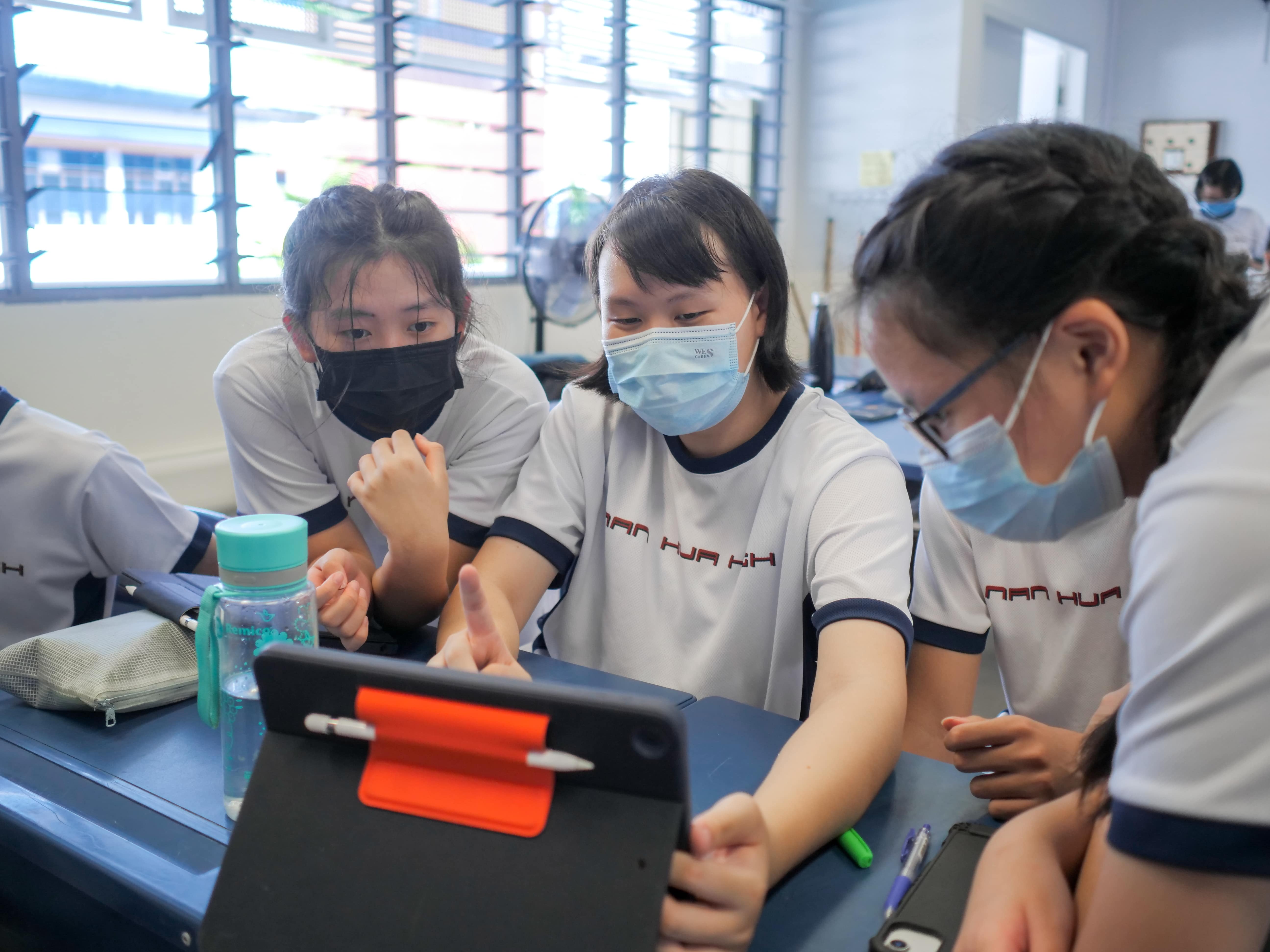 Three masked students in school uniforms gather around a tablet on a desk in a classroom.