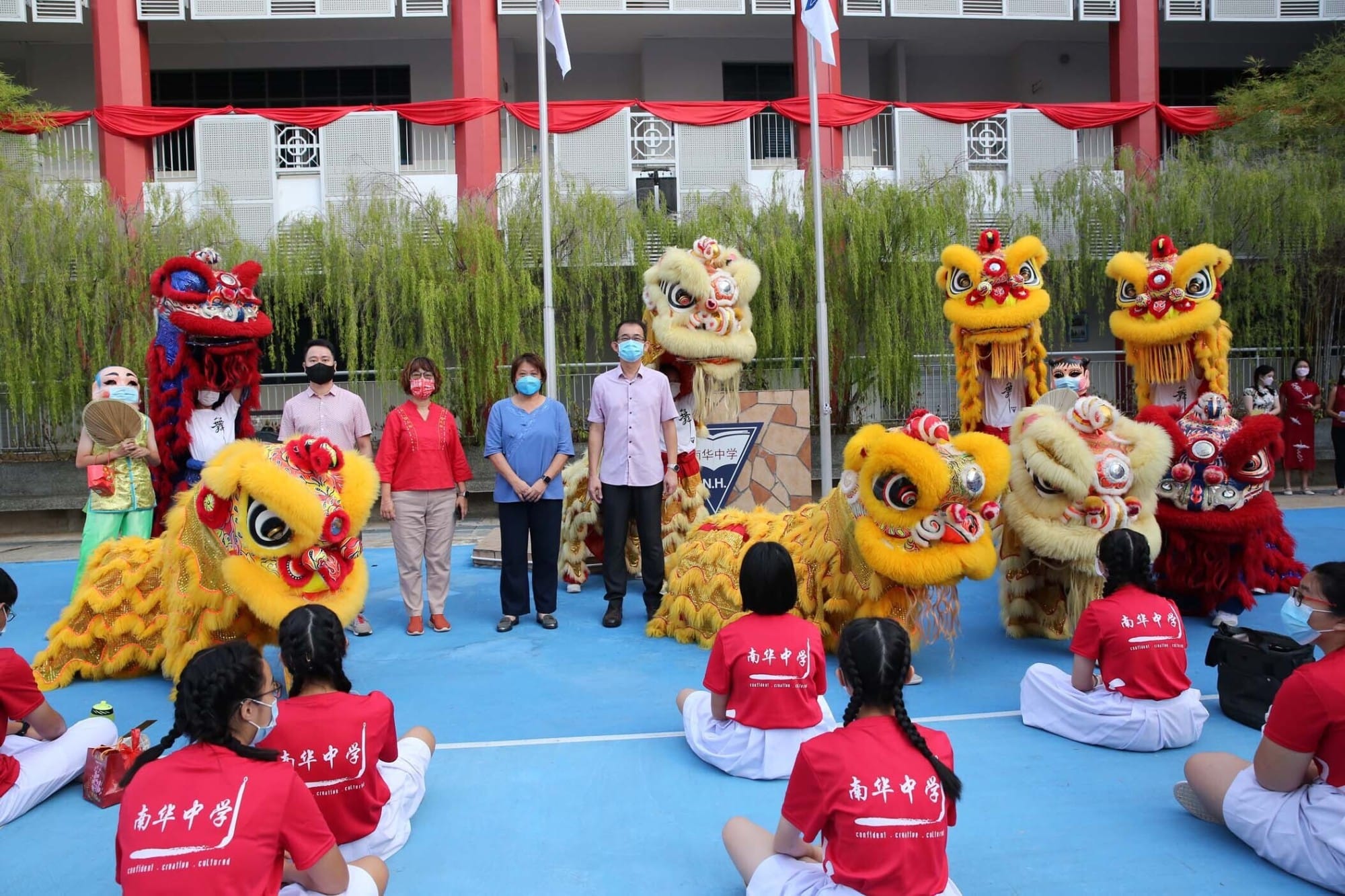 Students in red uniforms sit watching a colorful Chinese lion dance performance at a school courtyard with adults standing nearby.