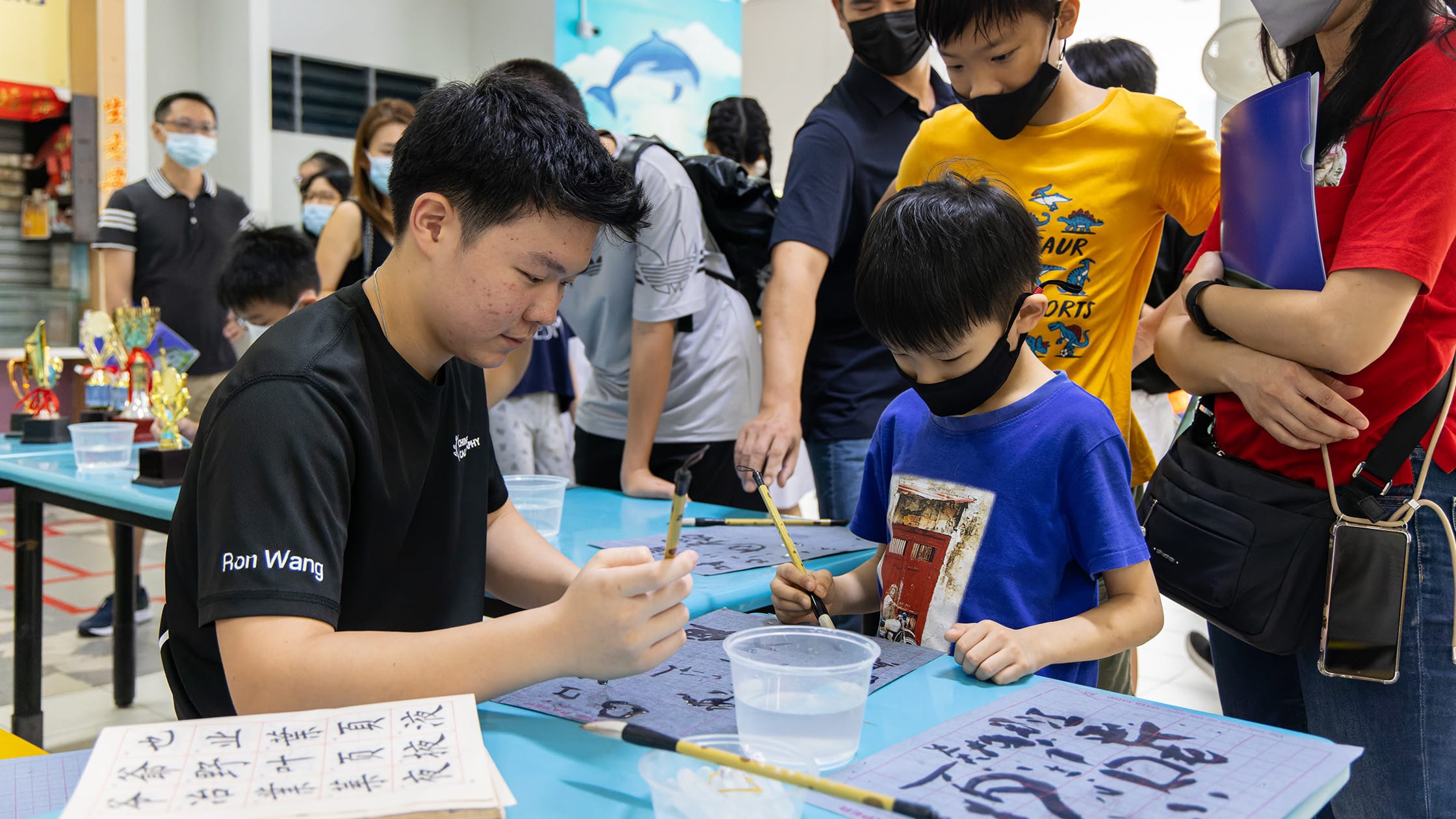 A teen teaches a young masked child Chinese calligraphy with brushes at a table during a school event.