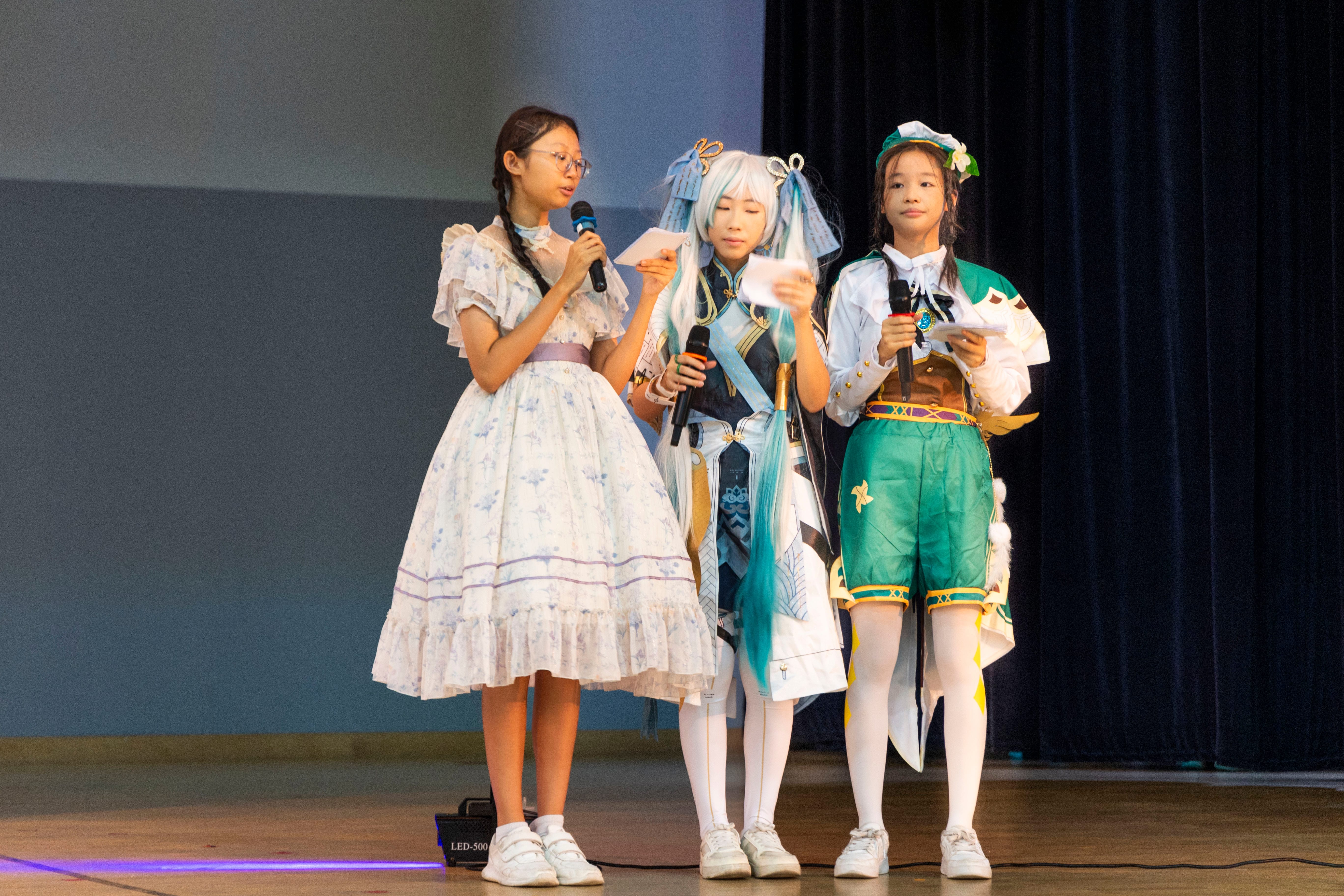 Three girls in cosplay costumes stand on a stage holding microphones and papers, performing or presenting.