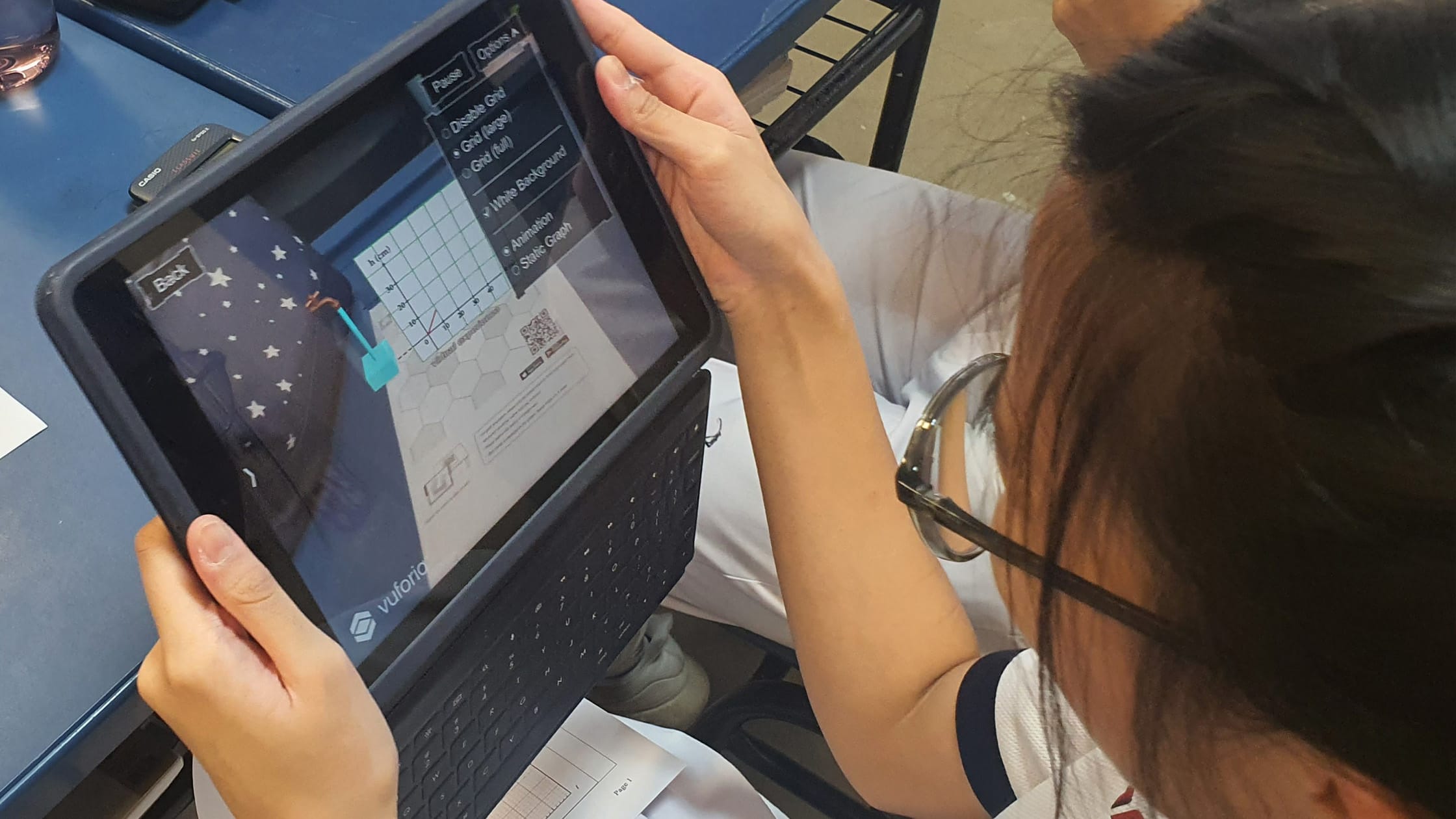 A student with glasses holds a tablet showing an AR math app with a graph, sitting at a blue desk in a classroom.